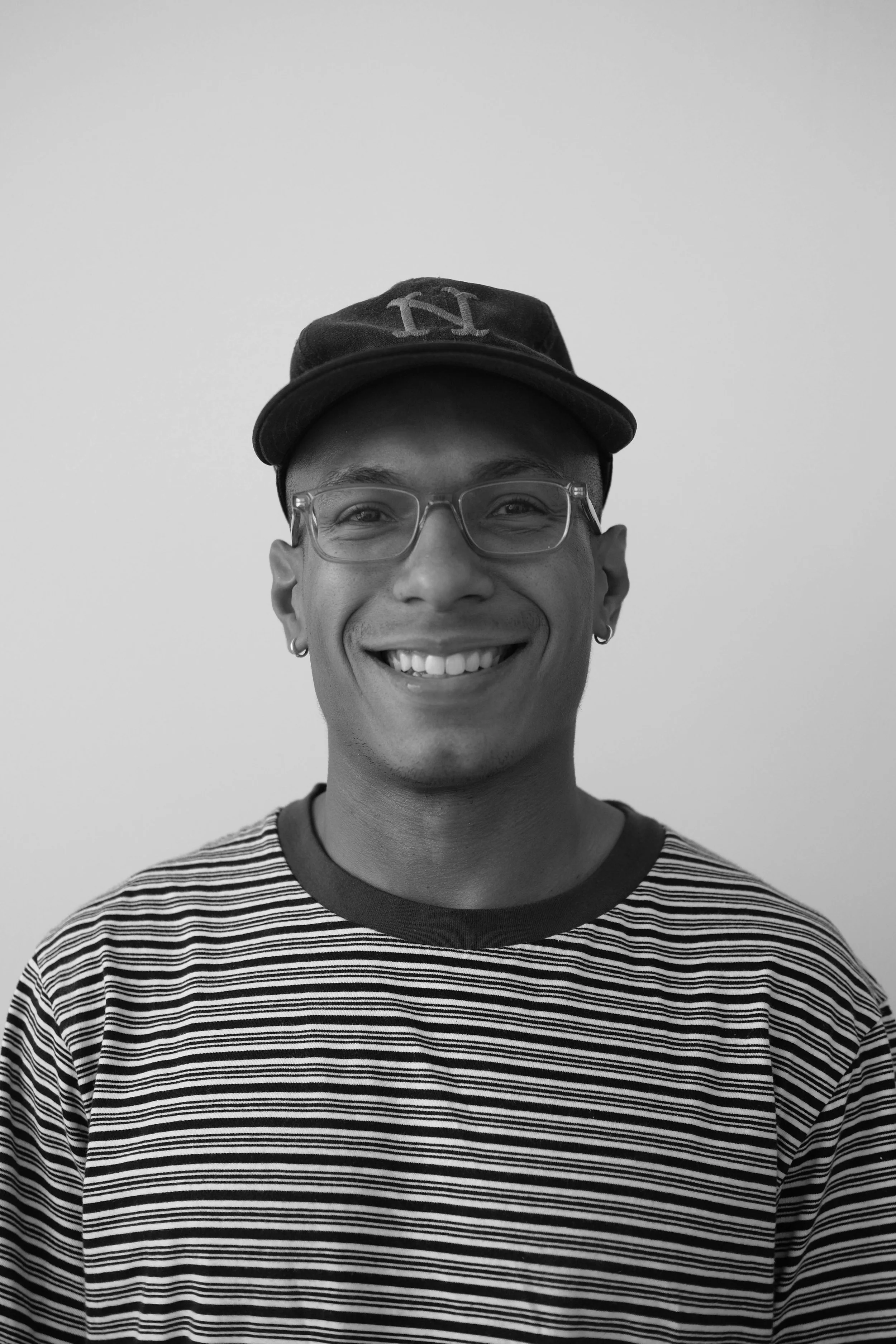 A smiling young man wearing glasses, a striped shirt, a cap with a letter N, and earrings, standing in front of a plain wall.