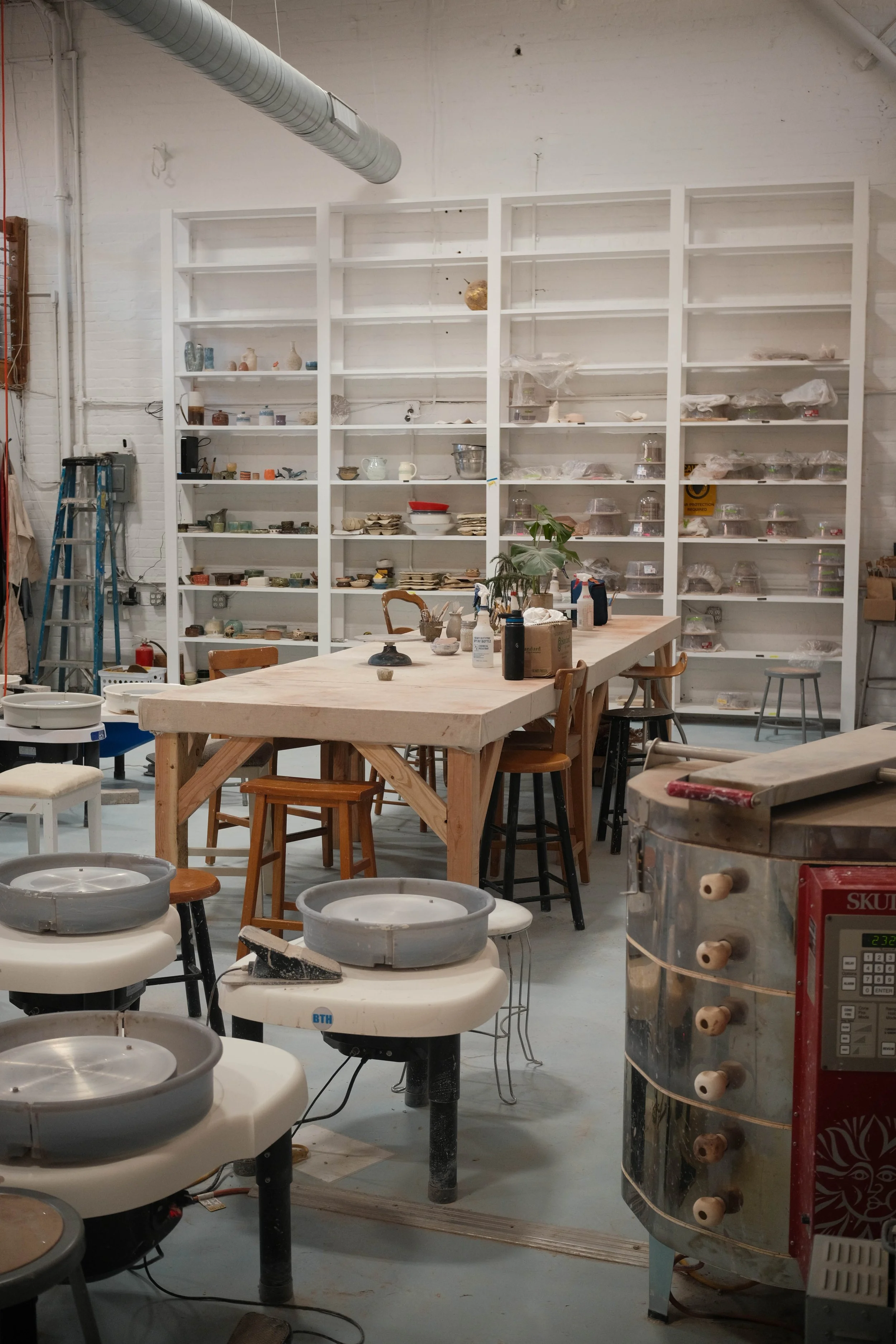 An art studio with pottery wheels, a large table, shelves with ceramics, and a machine in the foreground.