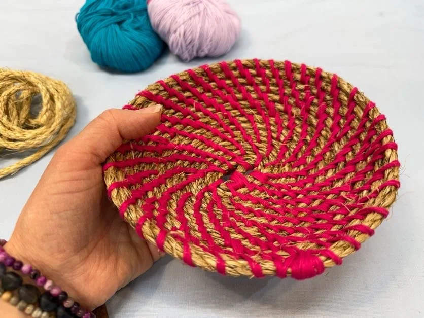 Person holding a woven basket with pink ribbon accents, with balls of yarn in teal and pink in the background.