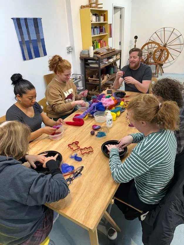 Group of six people sitting around a wooden table, working on felting projects with colorful wool and felting tools in a craft room.
