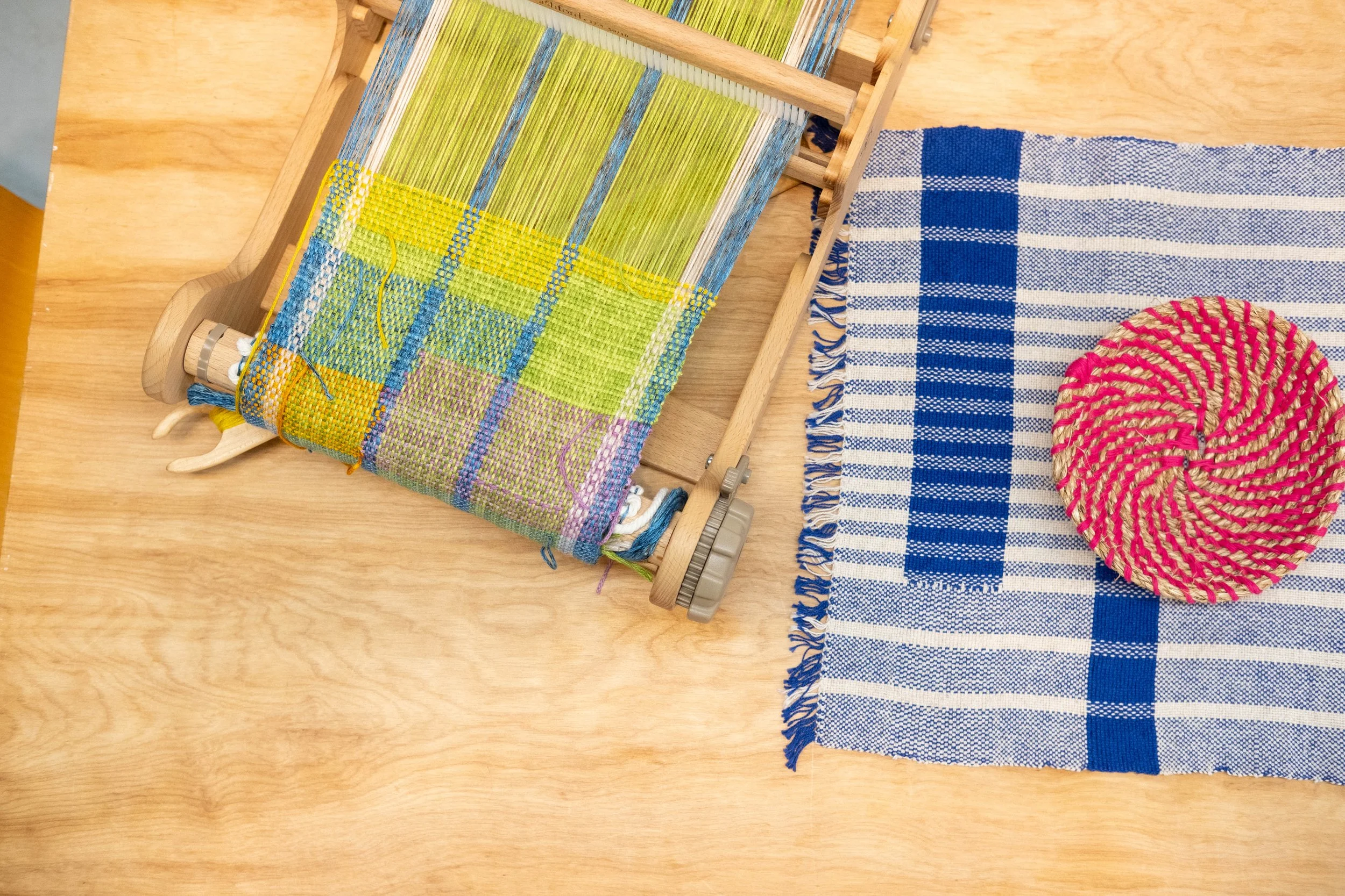 A wooden weaving loom with colorful woven fabric, a blue and white plaid fabric, and a round pink and beige woven coaster on a light wooden table.