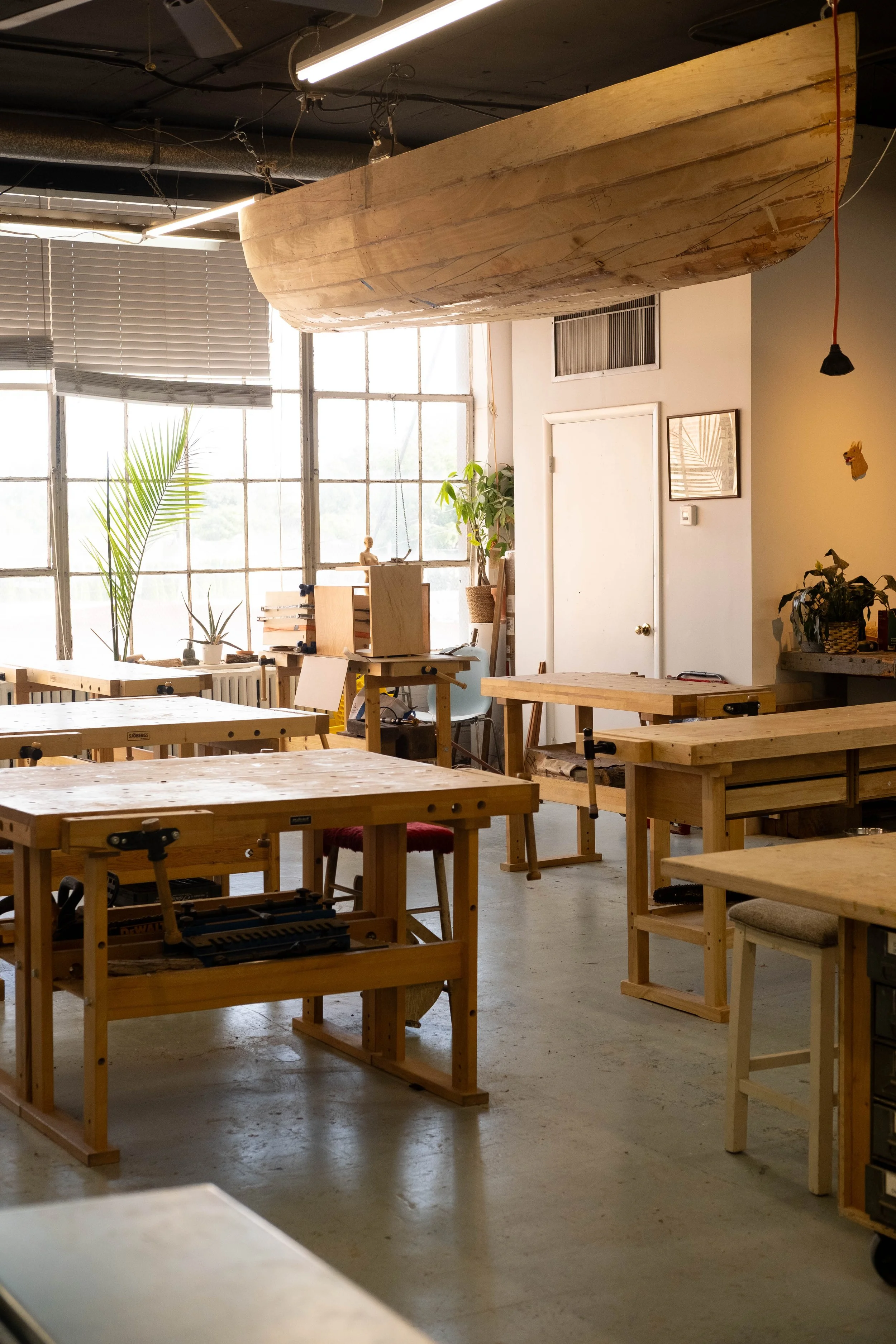Woodworking workshop with several workbenches and tools, large boat shadow above, large windows allowing natural light, plants, framed pictures on wall.
