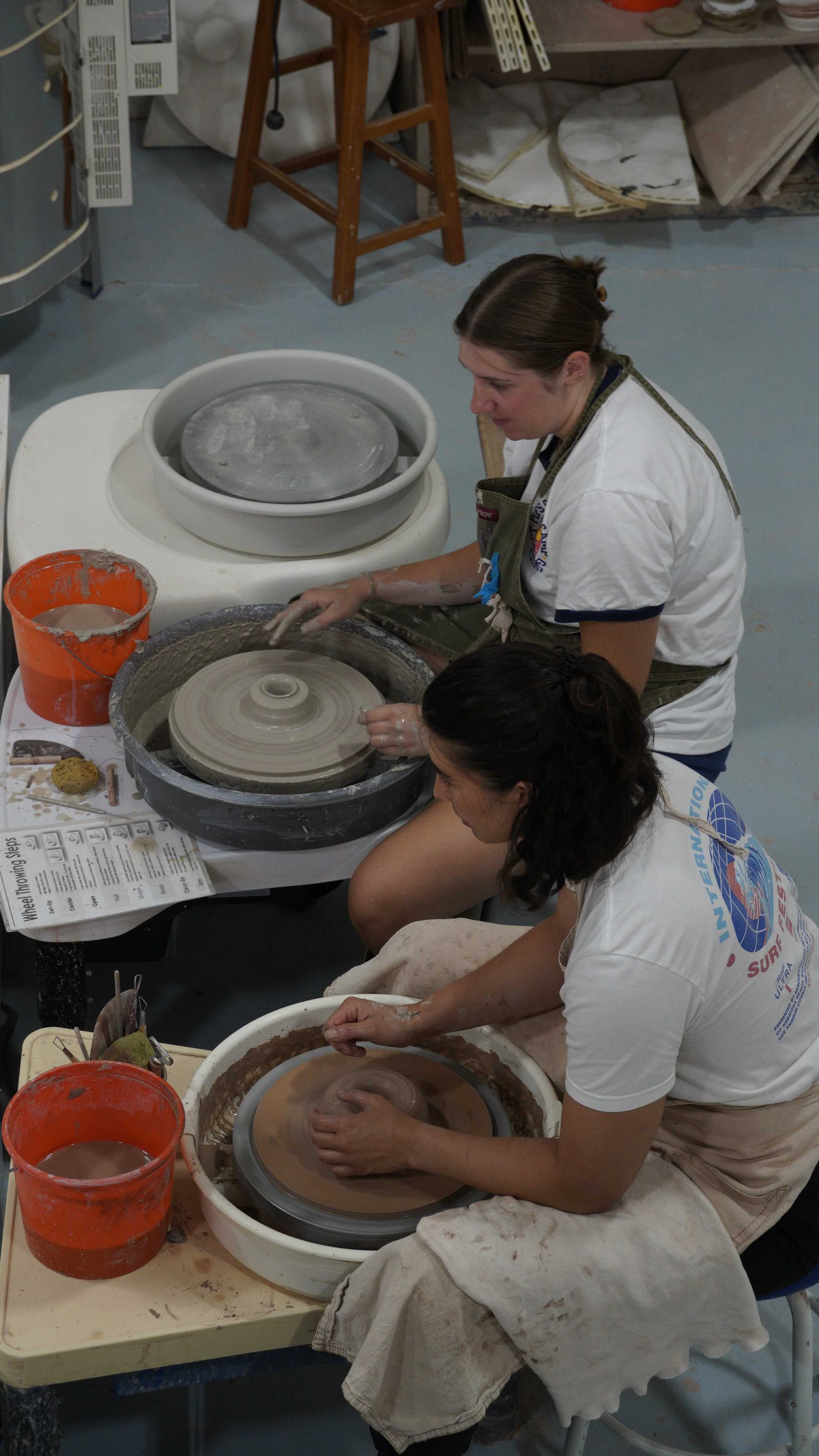 Two women working on pottery wheels shaping clay in a workshop, with supplies and tools on the table.