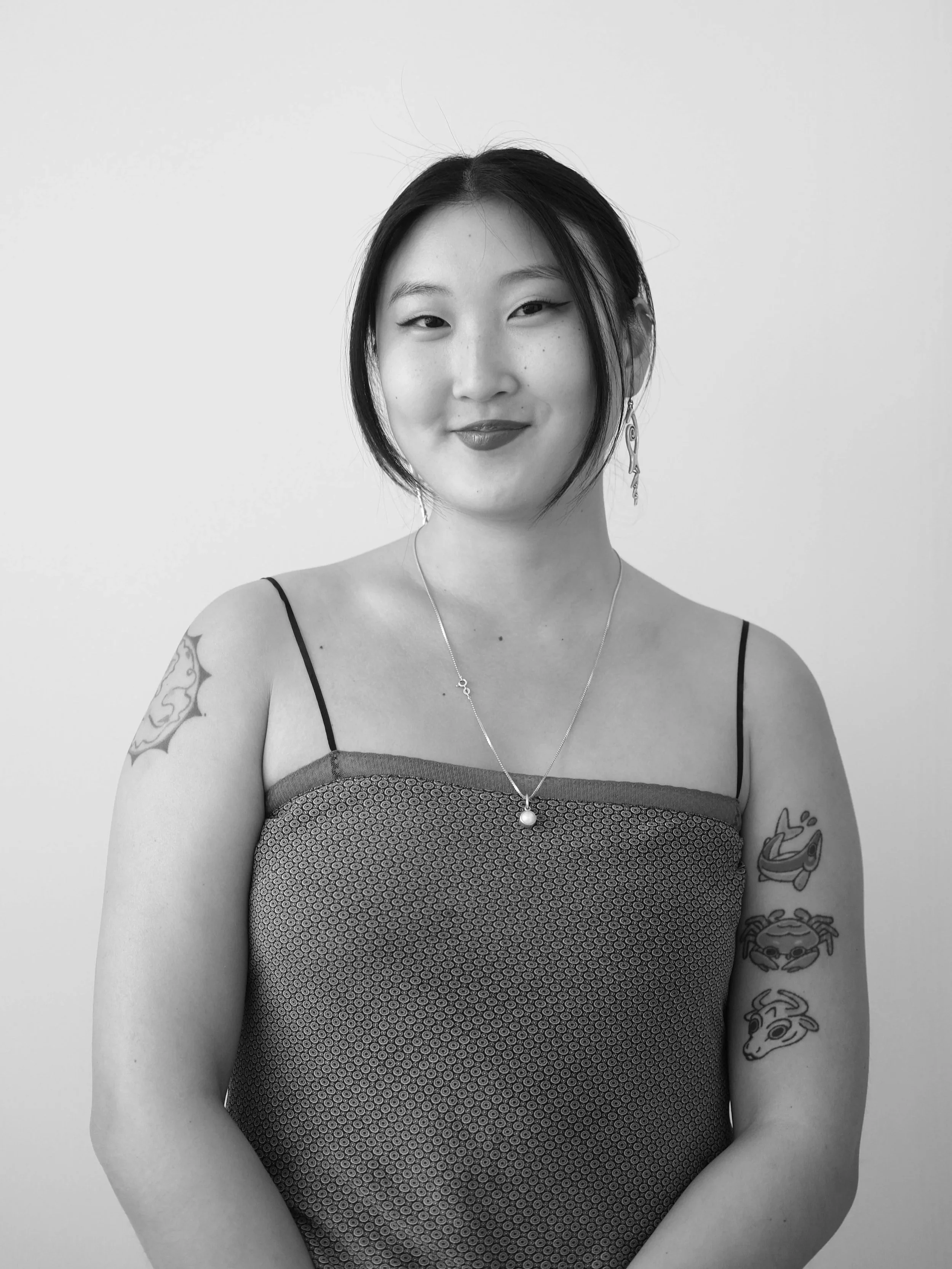 A black and white portrait of a woman with short hair, wearing a patterned tank top, jewelry, and tattoos on her arm.