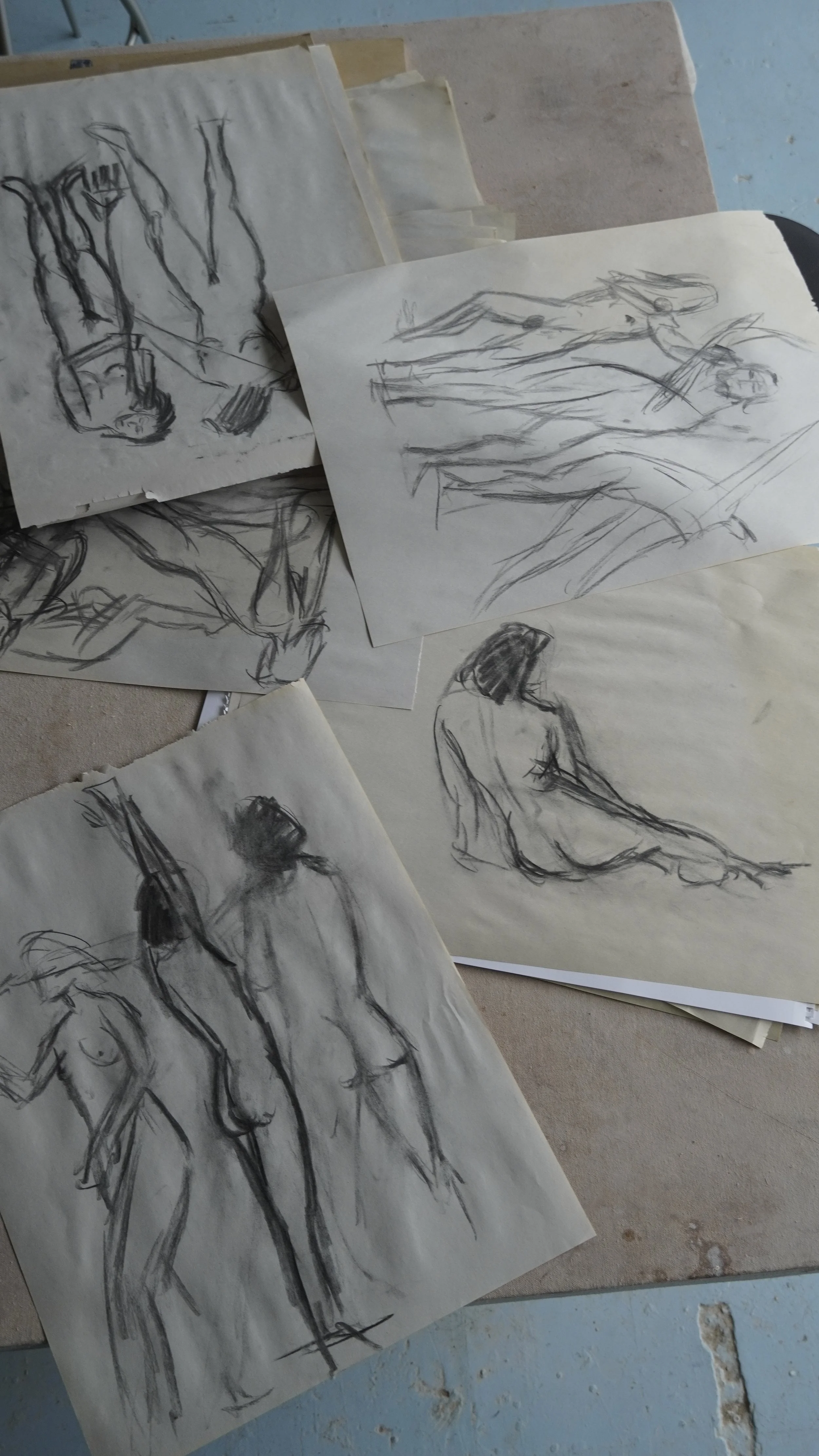 Collection of life drawing sketches of nude human figures, made with charcoal or pencil, on paper, arranged on a cluttered surface.