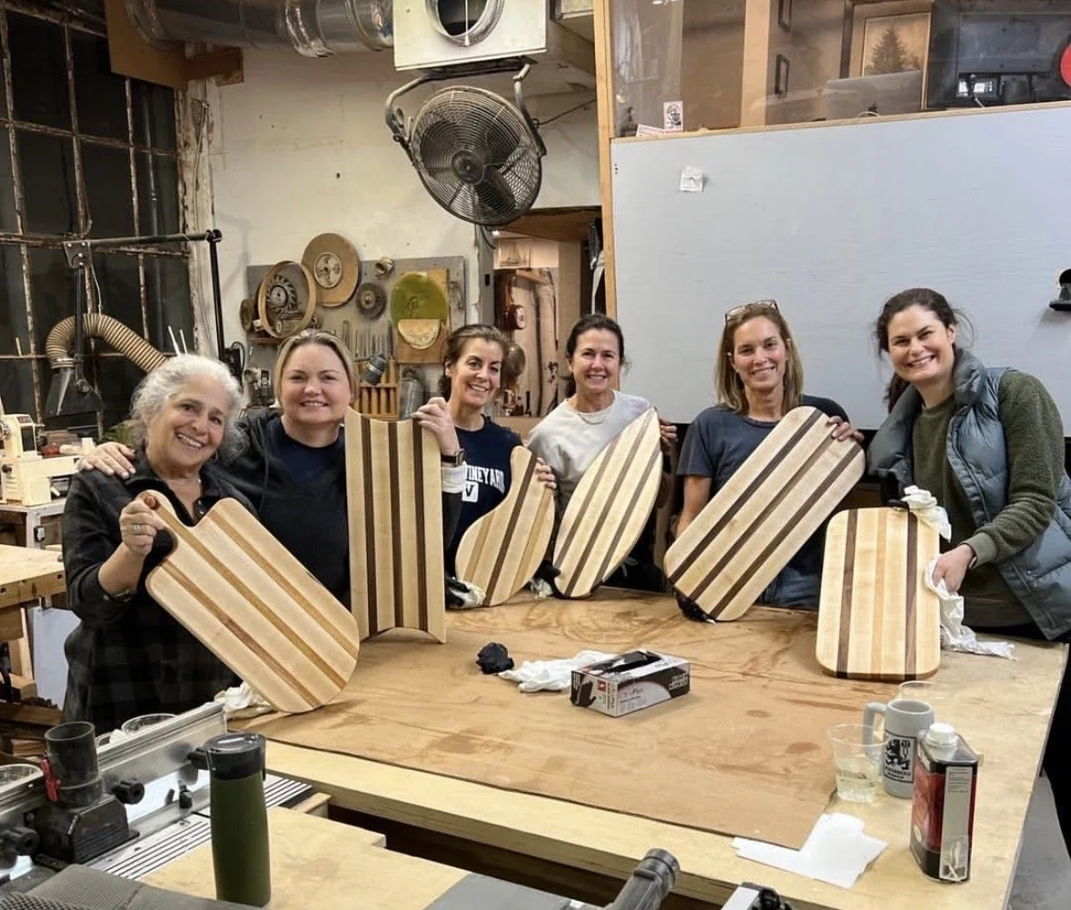 Six women standing behind a workbench in a woodworking shop, holding matching striped wooden cutting boards and smiling at the camera.