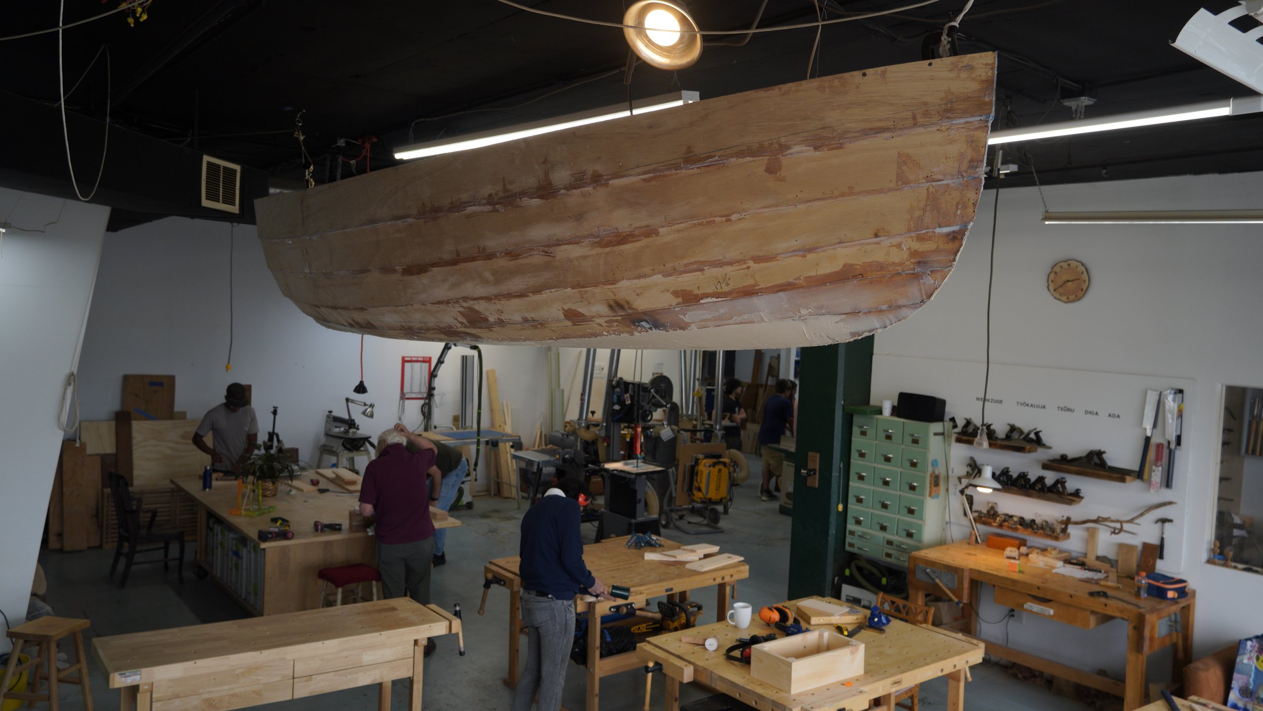 Woodworking workshop with people working on projects, tables, tools, and a large wooden boat hull hanging from the ceiling.