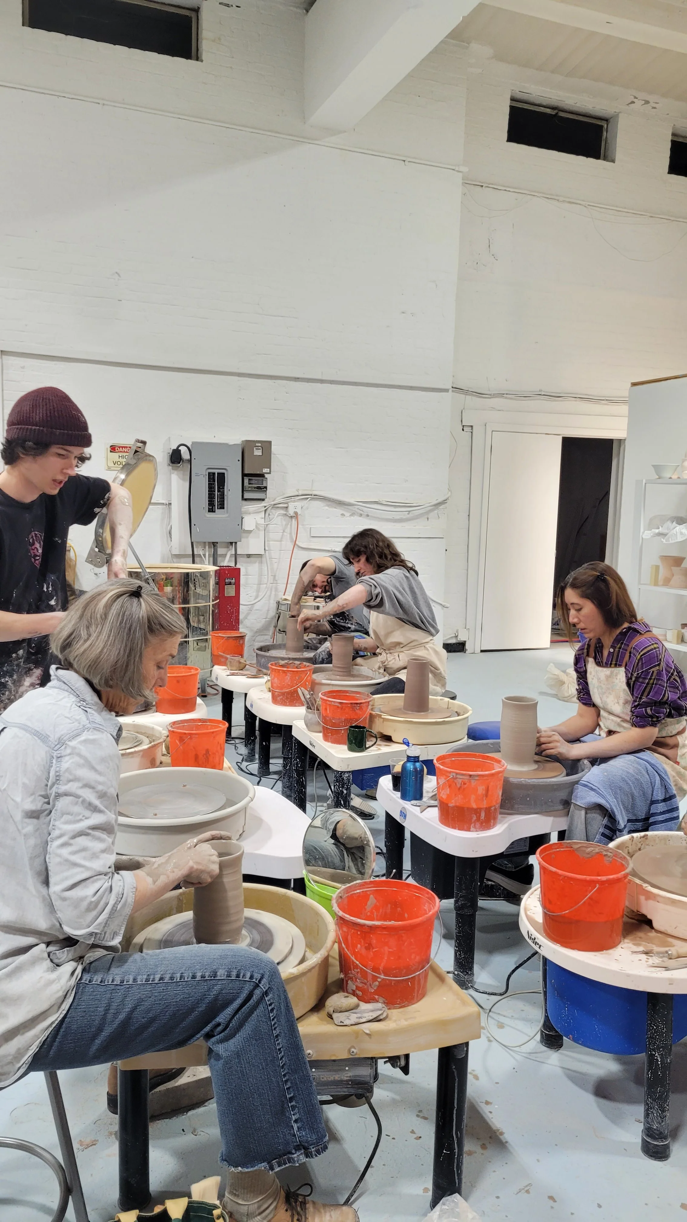 People participating in a pottery class, working on pottery wheels in a spacious room with white walls and high ceiling.
