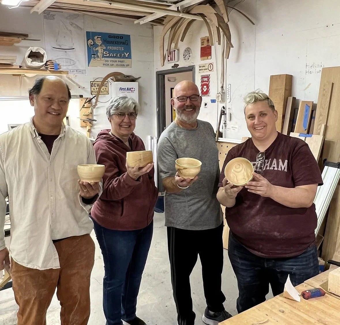 Four people standing inside a woodworking shop, each holding a wooden bowl and smiling at the camera.