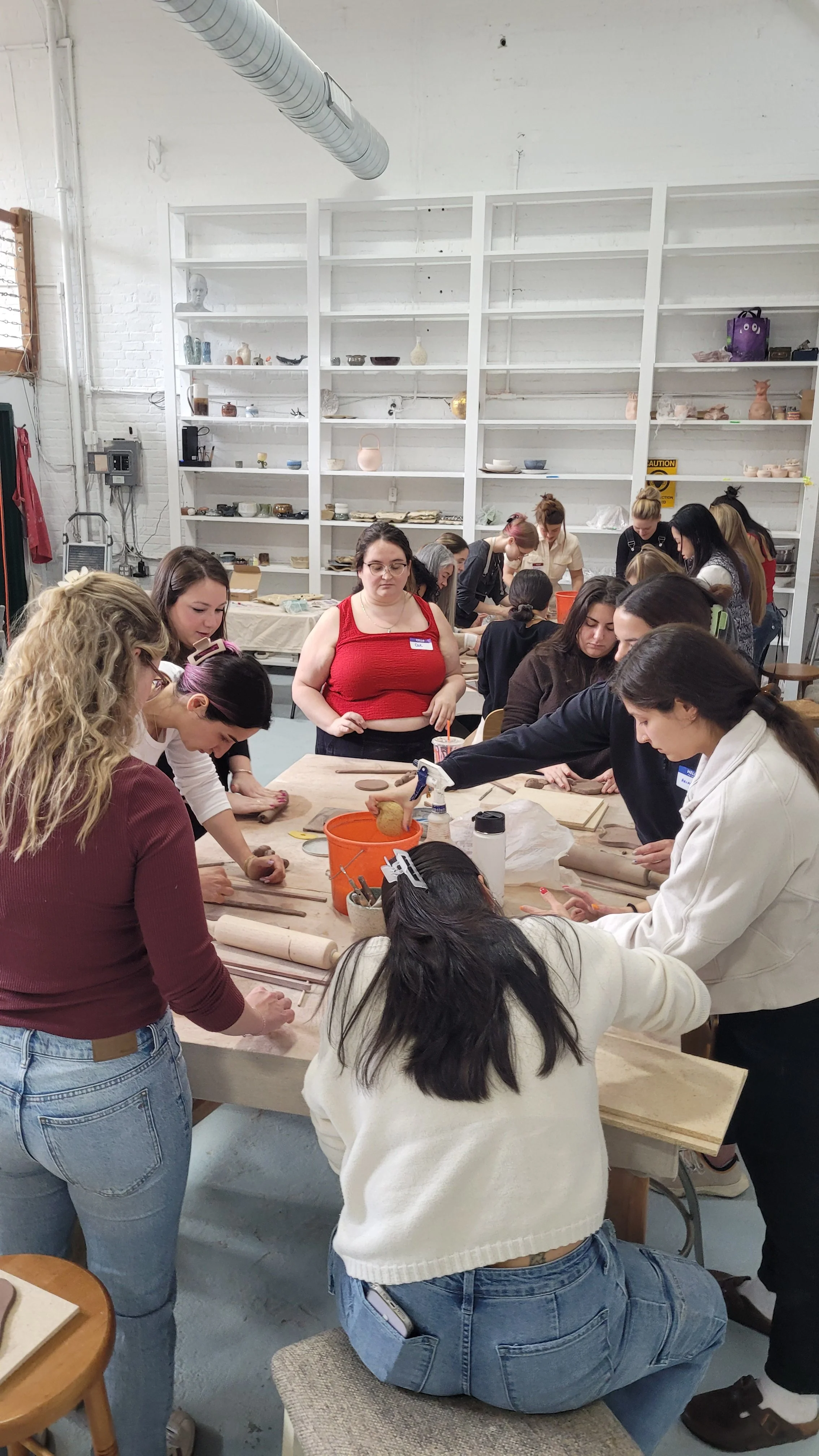 Group of women participating in a pottery or ceramics workshop in a studio with shelves of pottery on the wall.