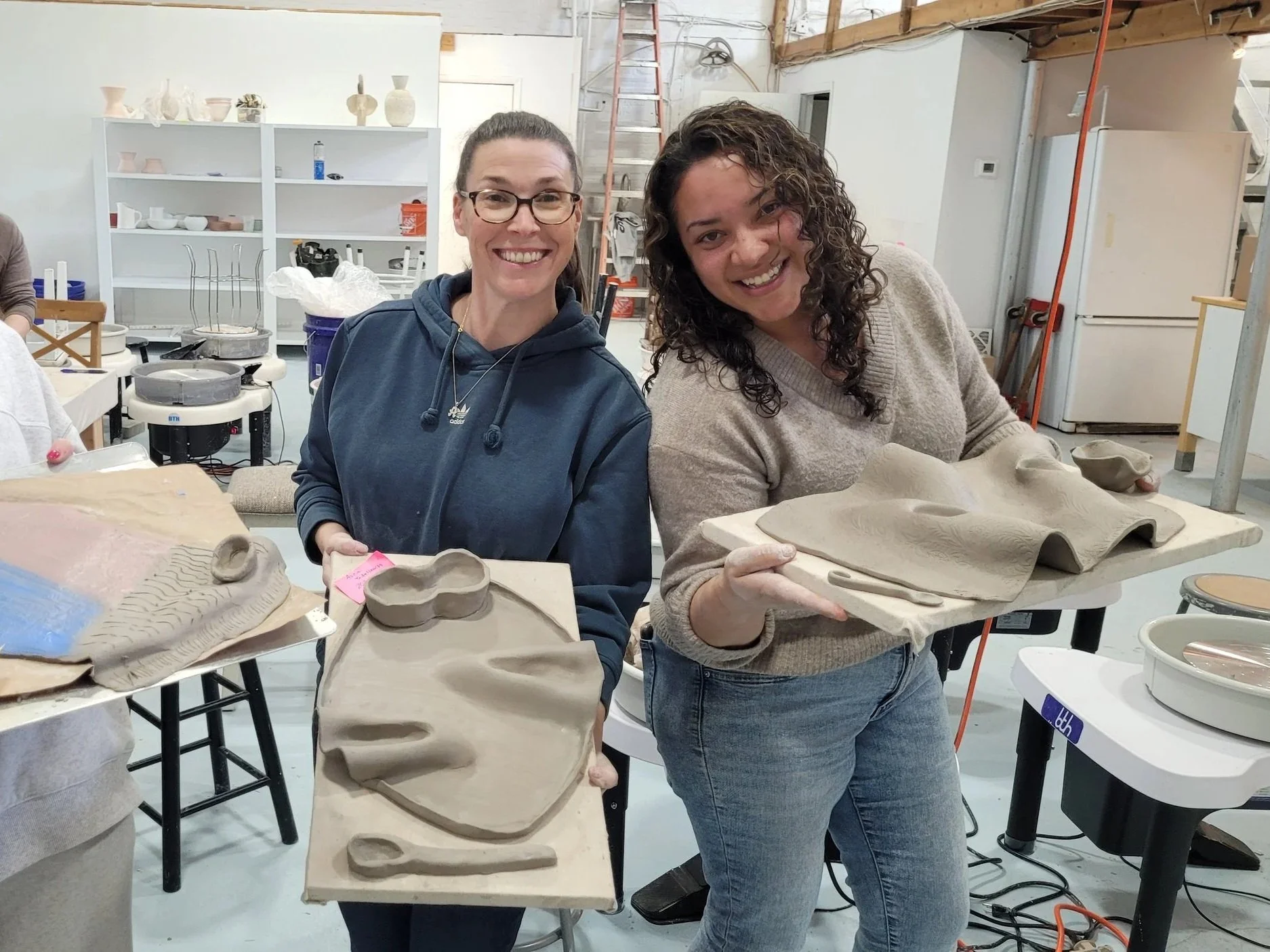 Two women smiling while holding pottery pieces in a pottery studio. Shelves with pottery and equipment are visible in the background.