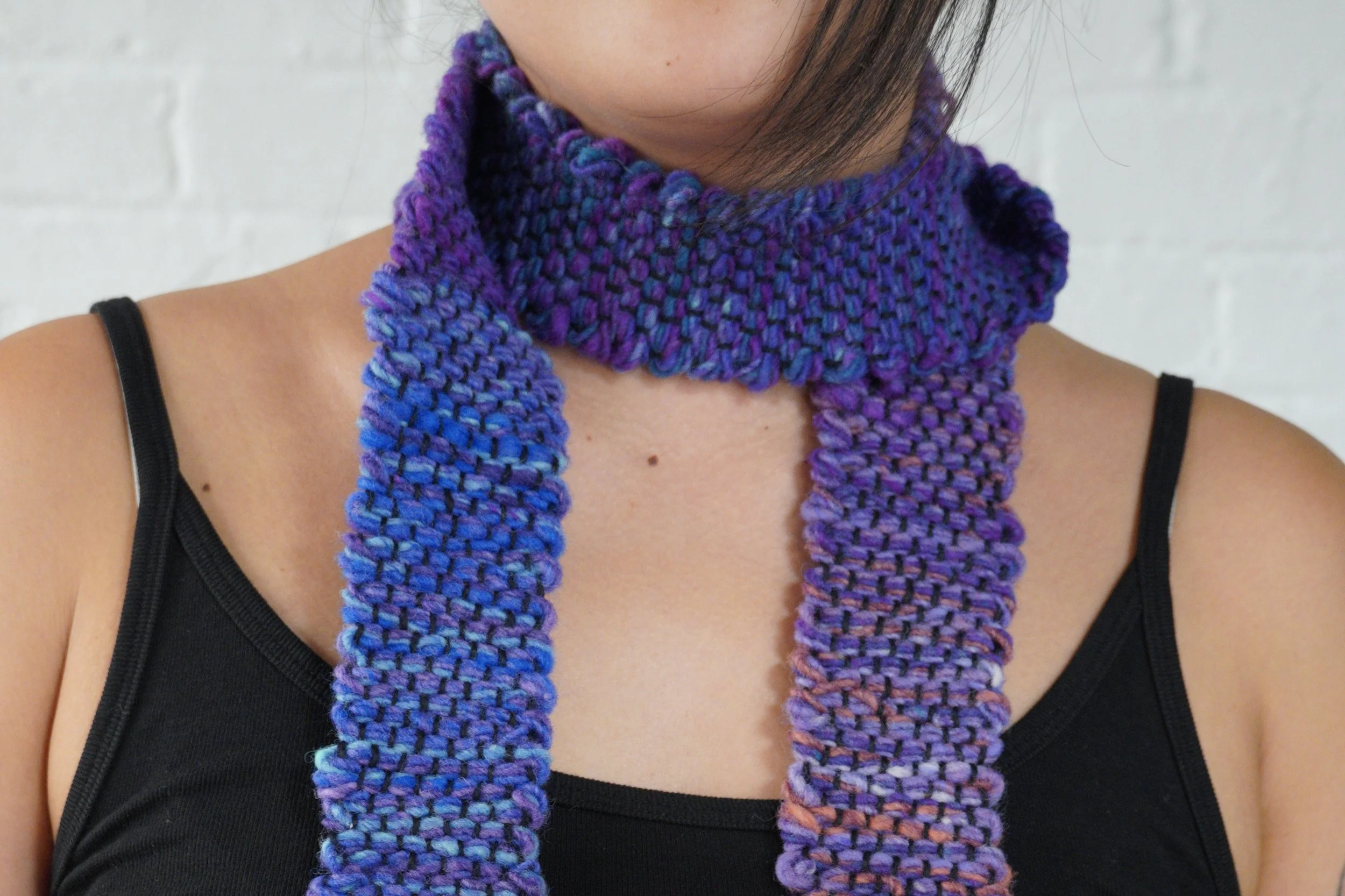 Person wearing a black tank top and multicolored purple, blue, and gray knitted scarf.