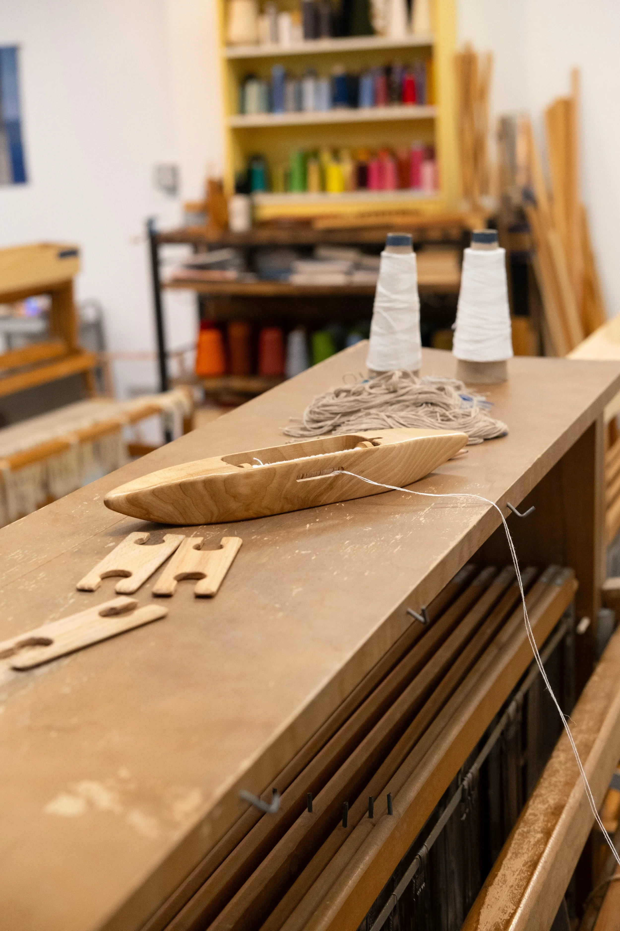 Two spools of white thread on a wooden surface in a craft or woodworking workshop, with a wooden boat-shaped object and some letter-shaped cutouts nearby. In the background, shelves with colorful spools of thread and various woodworking supplies are 
