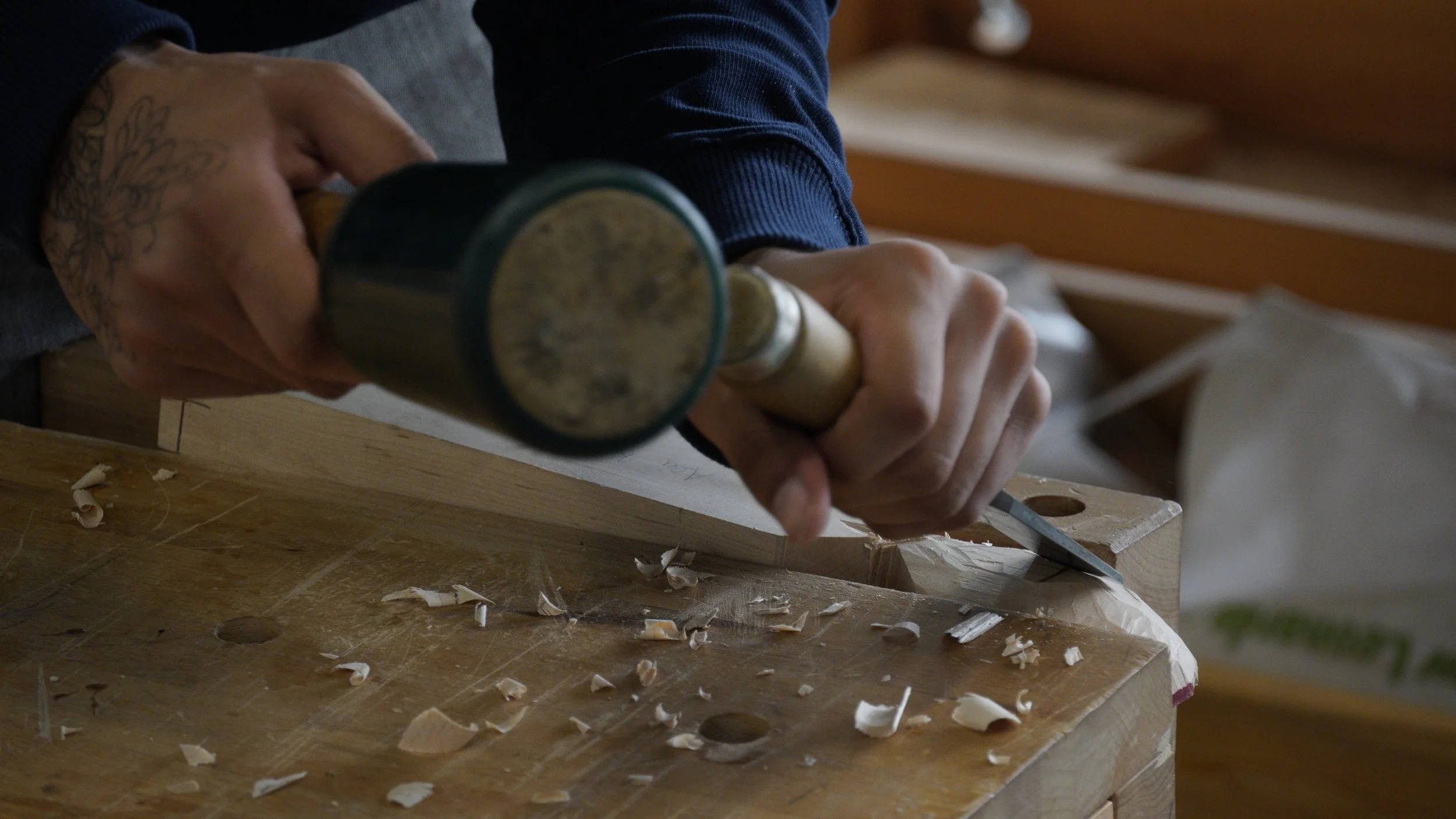 Person using a hammer to carve or shape wood on a workbench, with wood shavings scattered around.