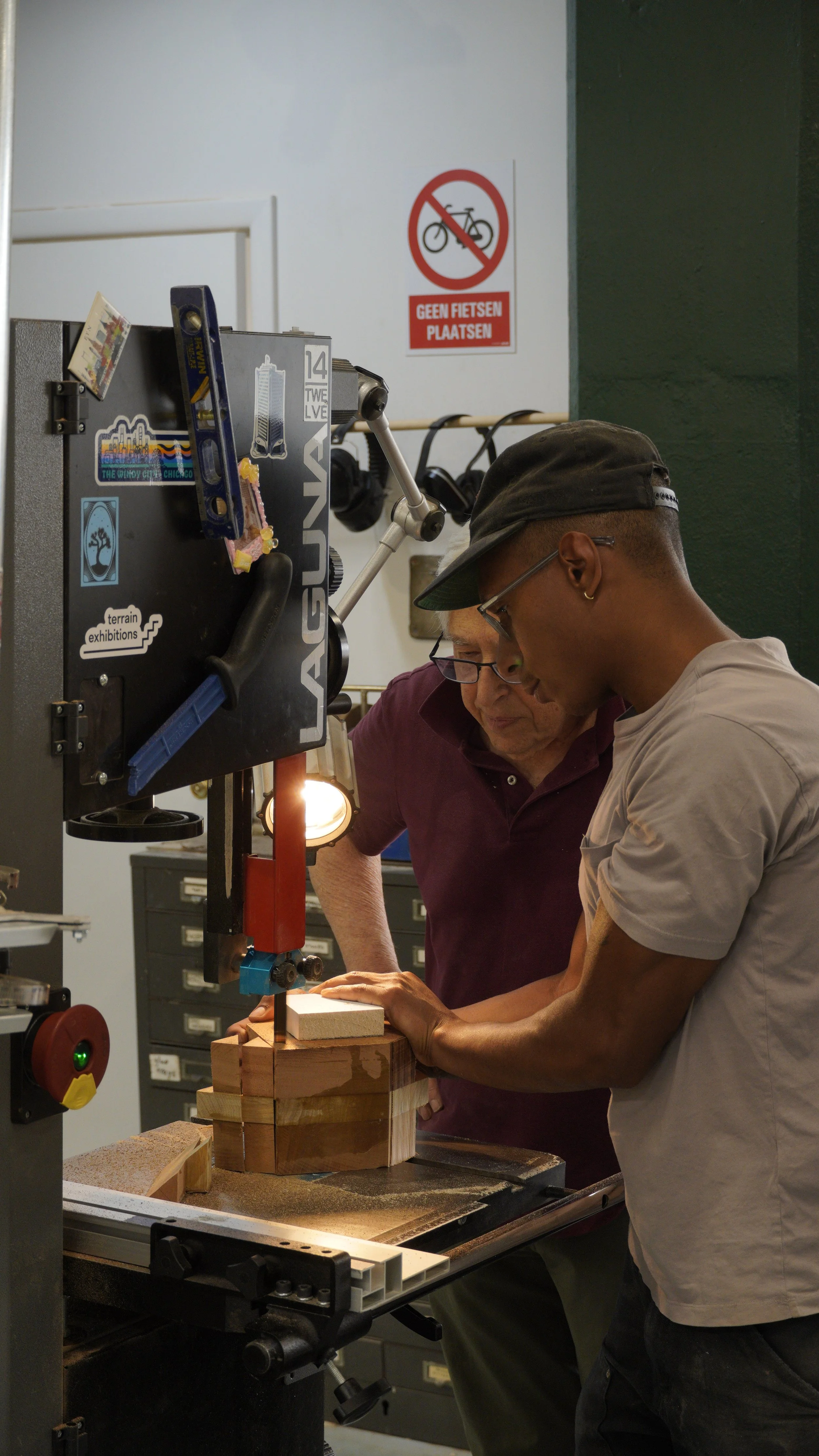 Two men working with a bandsaw in a workshop, cutting a wooden piece, with a 'No Bicycles' sign on the wall behind them.