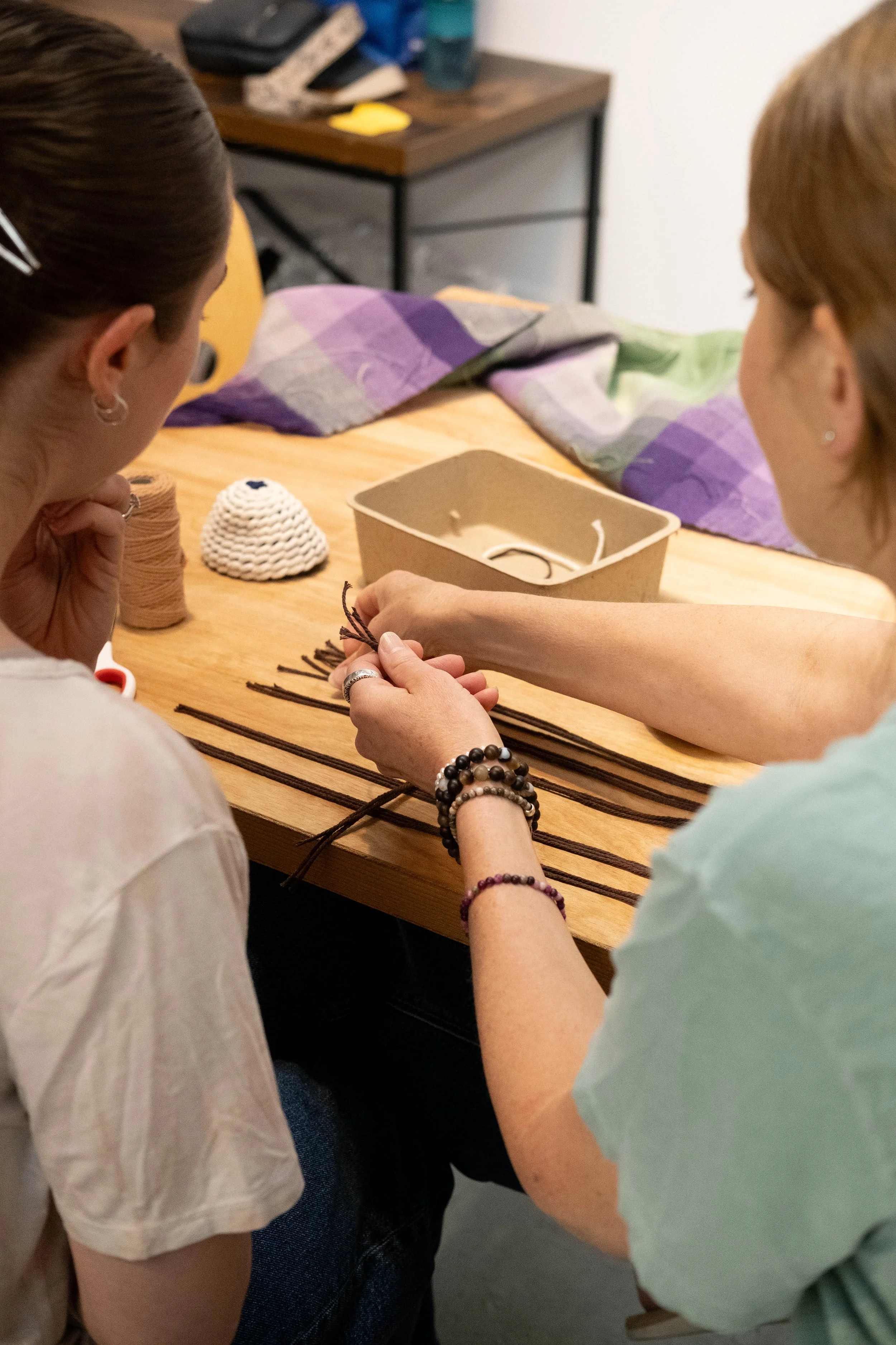 Two women are sitting at a wooden table, making rope with twisted strands. There are spools of thread, crochet pieces, and a cardboard box on the table. One woman has bracelets on her wrist, and the other woman is holding the rope. The background sho