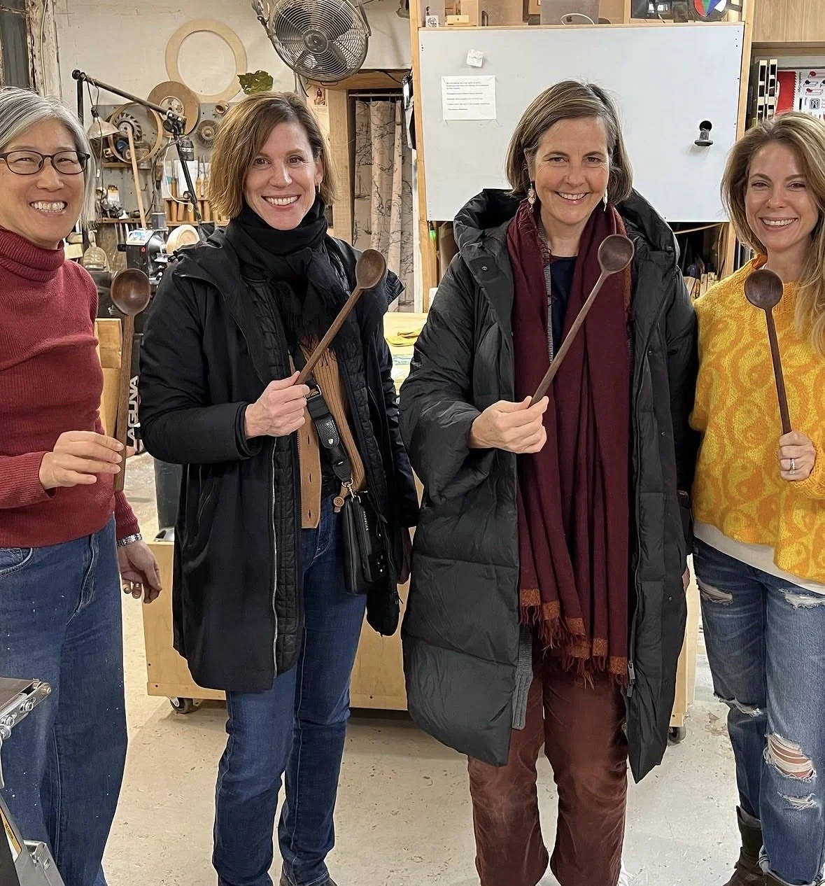 Four women standing in a workshop holding kitchen spoons, smiling at the camera.