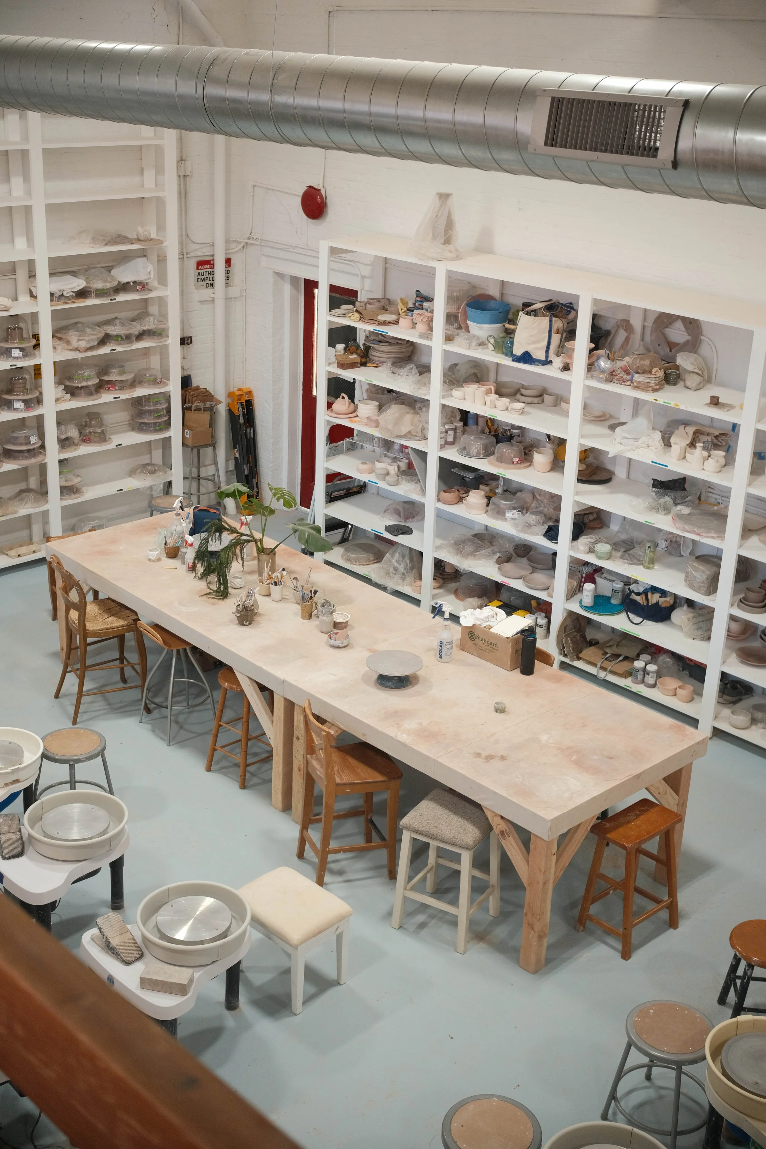 A ceramics studio with shelves filled with unfinished pottery and clay objects, a long worktable with tools, and chairs around it.