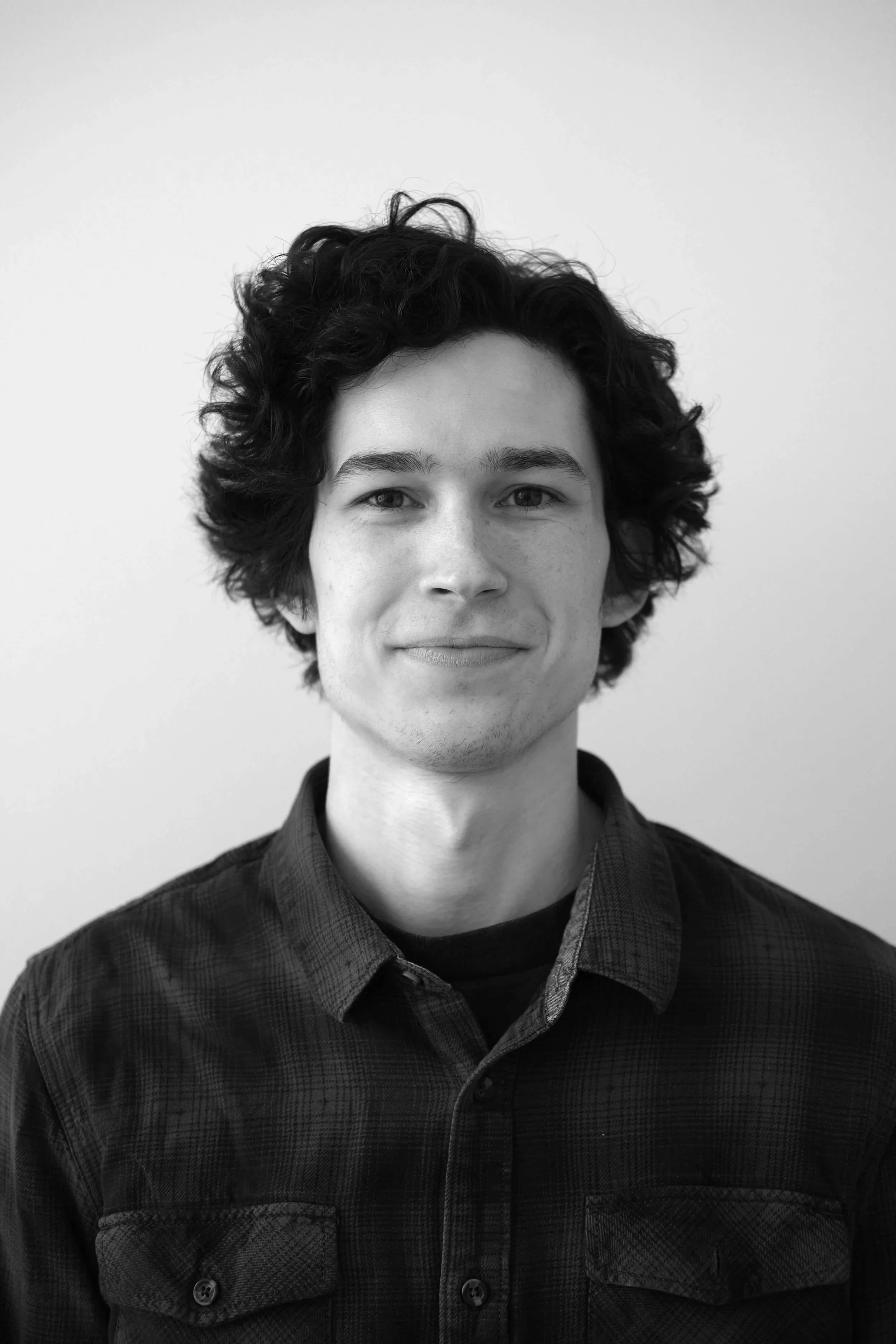 A young man with curly dark hair, light skin, wearing a dark plaid button-up shirt, smiling slightly, against a plain light-colored background.