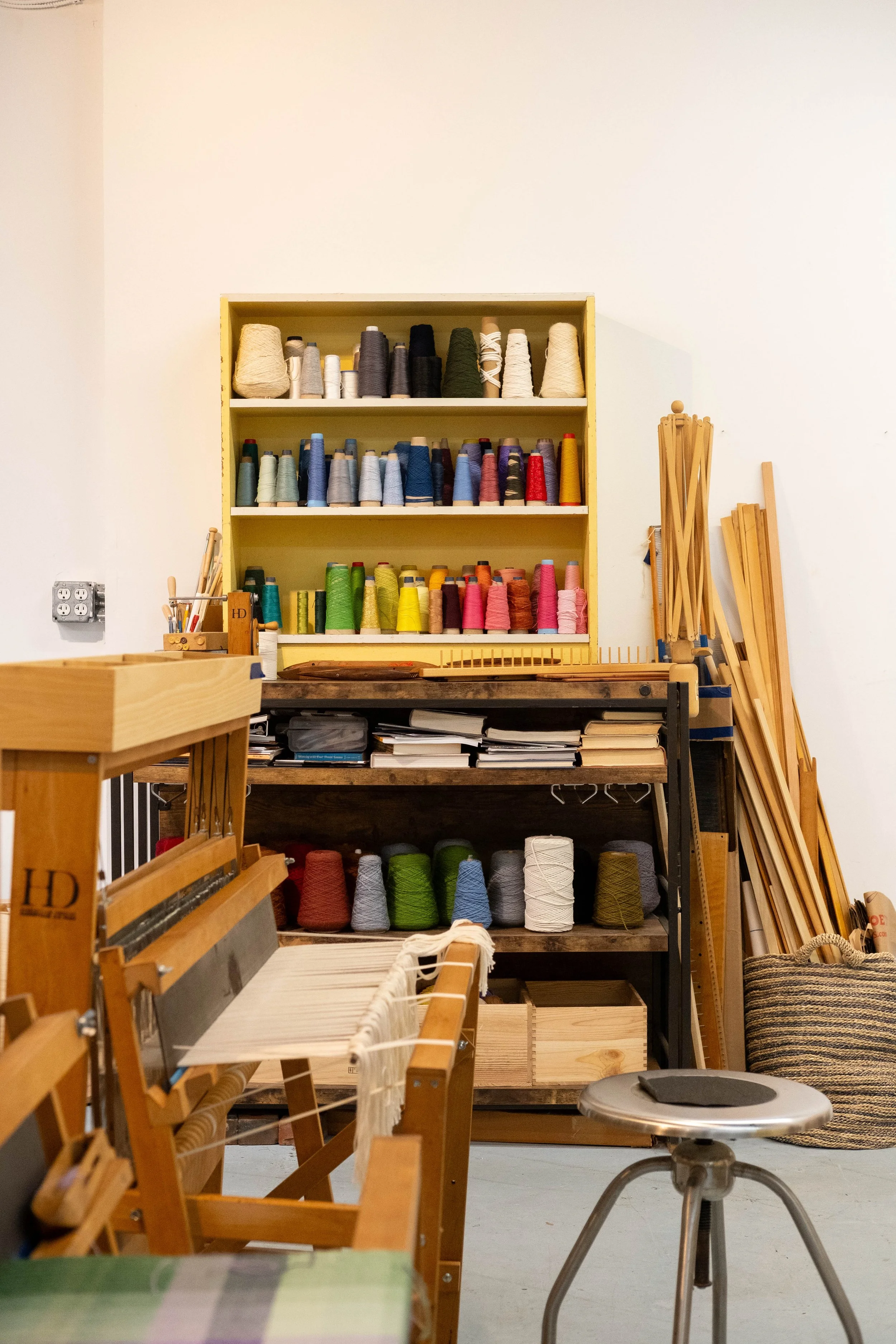 A shelving unit with multiple rows of colorful yarn spools, yarn weaving loom in the foreground, and wooden weaving frame on the right side in a craft workshop or studio.