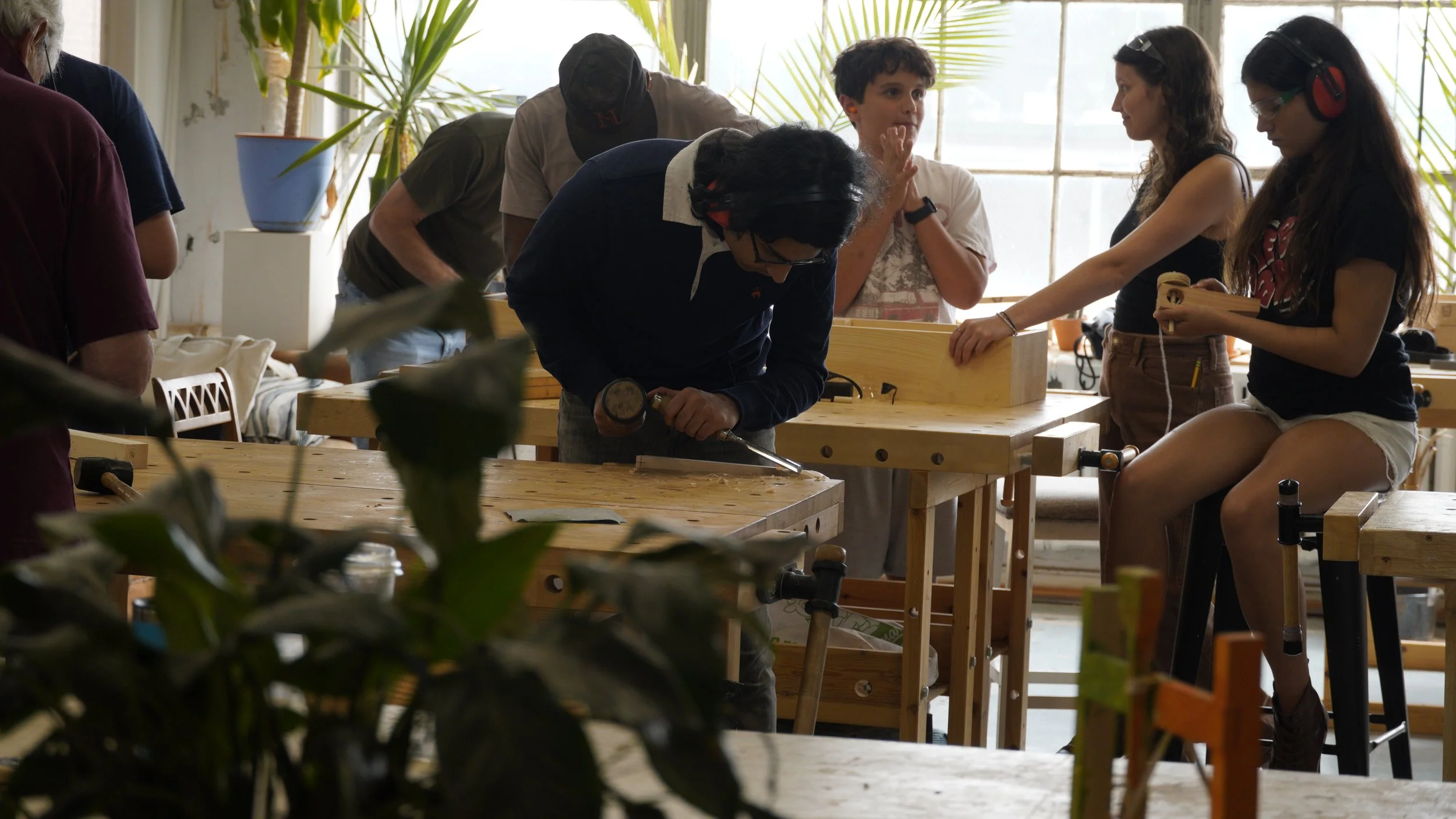 People engaging in woodworking activities in a bright, plant-filled workshop, with some wearing protective ear gear and using tools on wooden tables.