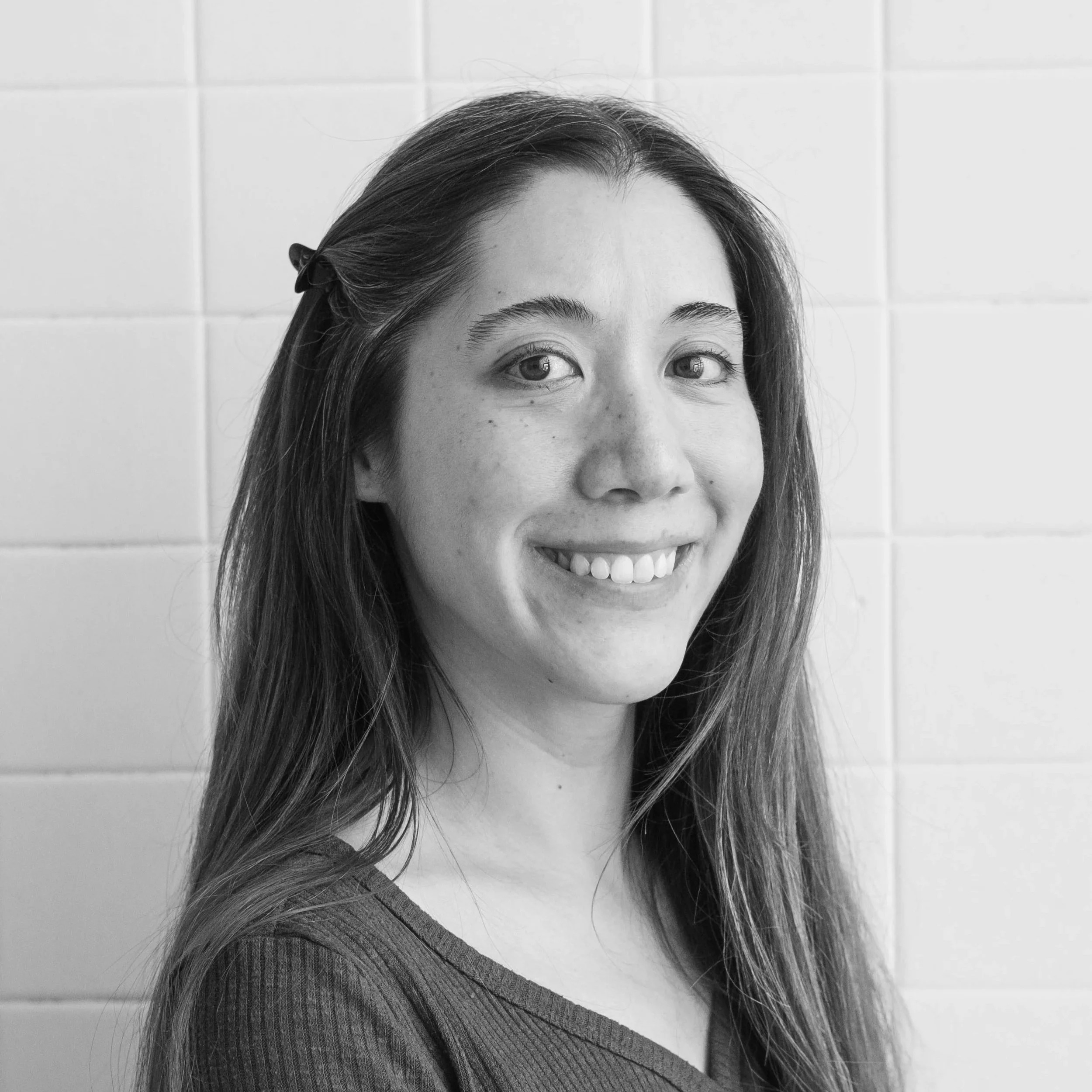 Black and white photo of a smiling woman with long hair, wearing a textured shirt, against a tiled wall background.