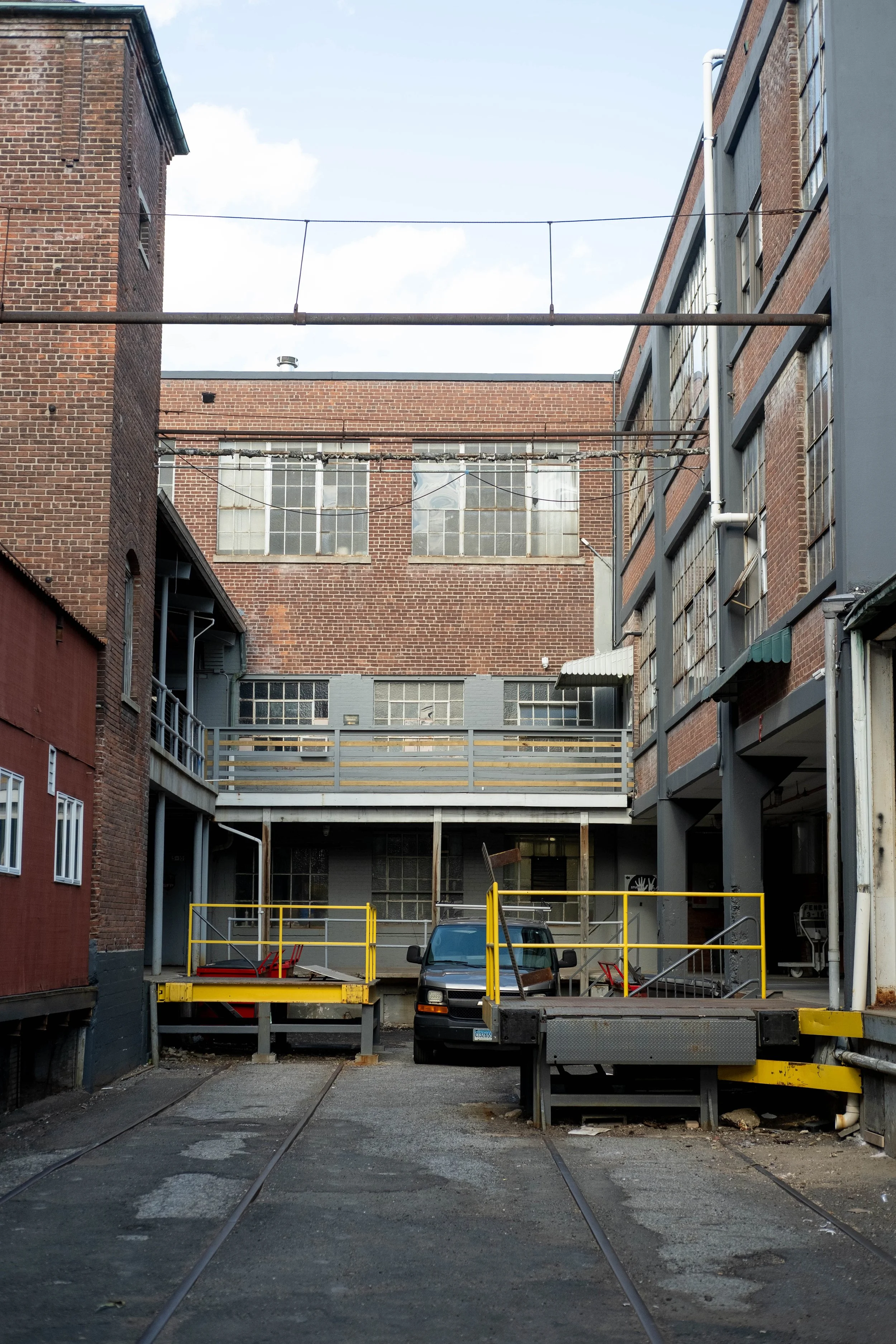 An alleyway surrounded by brick and gray buildings, with two sets of train tracks running through it. A silver pickup truck is parked, and there is a yellow barrier gate at the entrance. Overhead, there are electrical wires and a sky with some clouds.