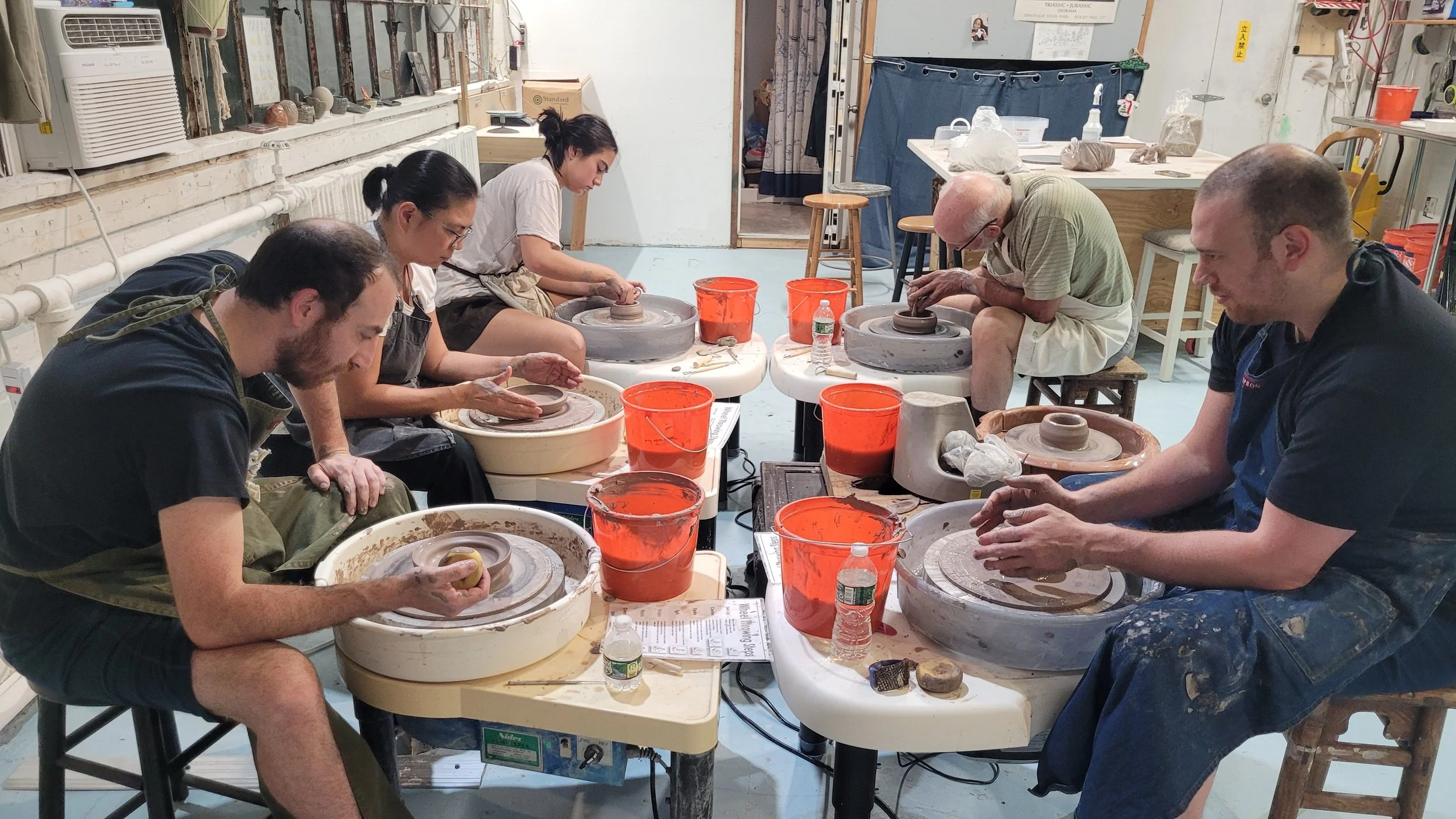 People participating in a pottery class, sitting at pottery wheels shaping clay, with buckets of water and pottery tools on the tables in a studio.