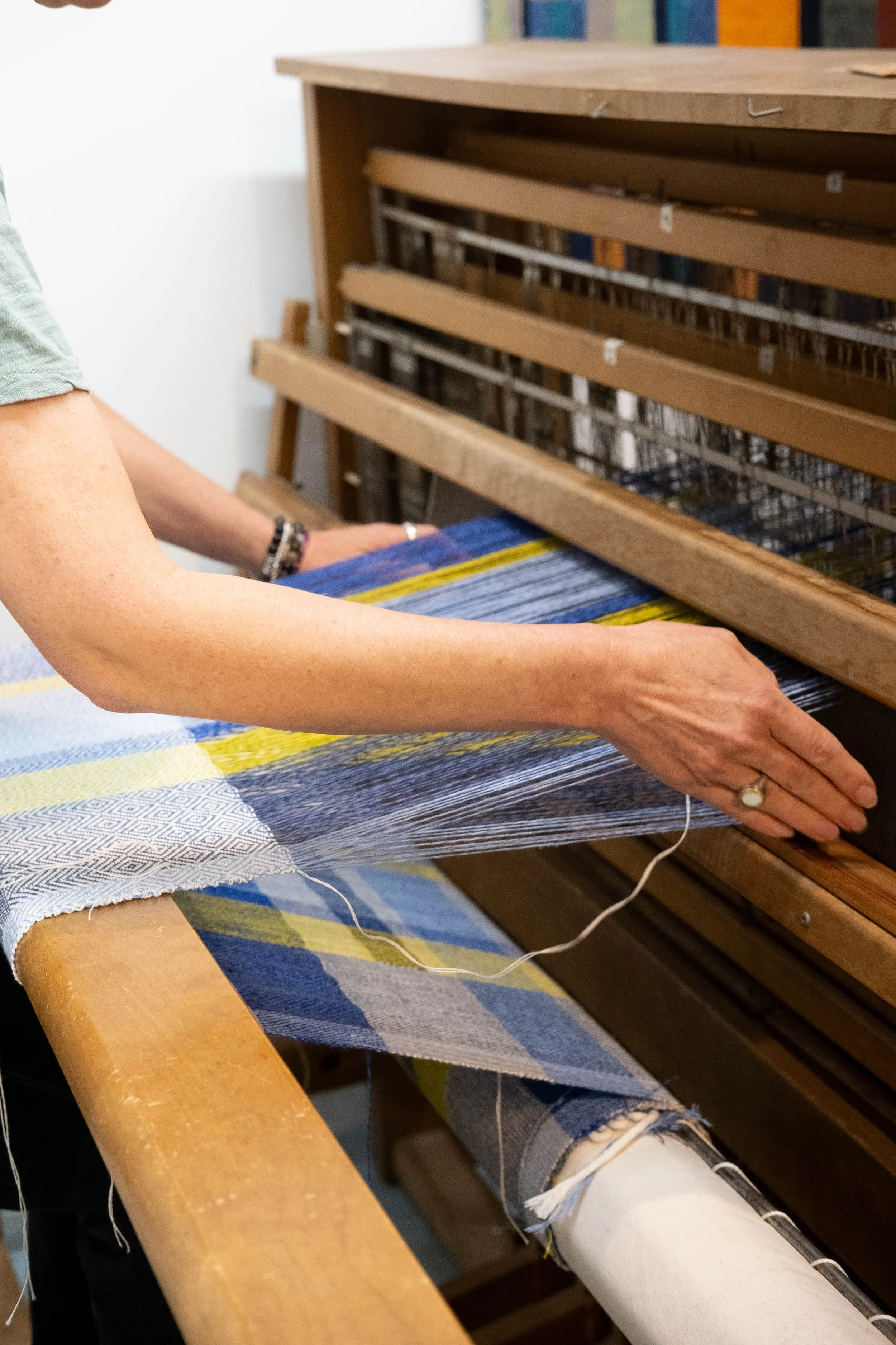 Person weaving on a loom with colorful fabric, wearing a light green shirt and jewelry.