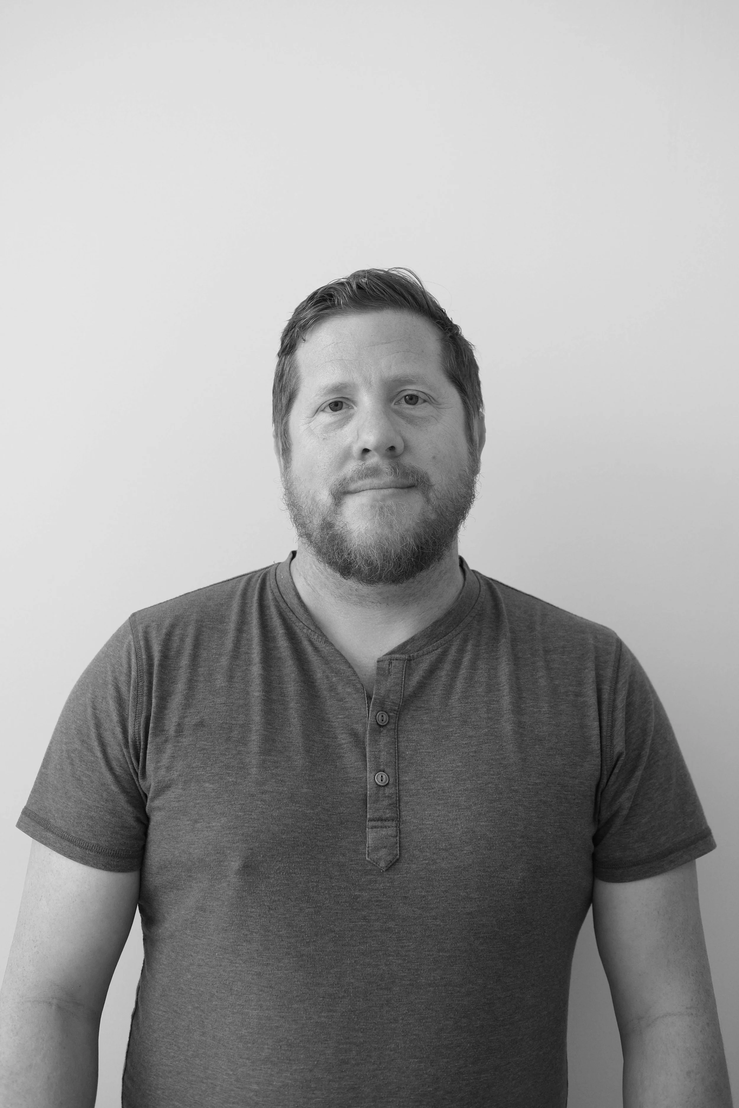 Black and white photo of a man with short hair and a beard, wearing a short-sleeved, buttoned shirt, standing against a plain wall.