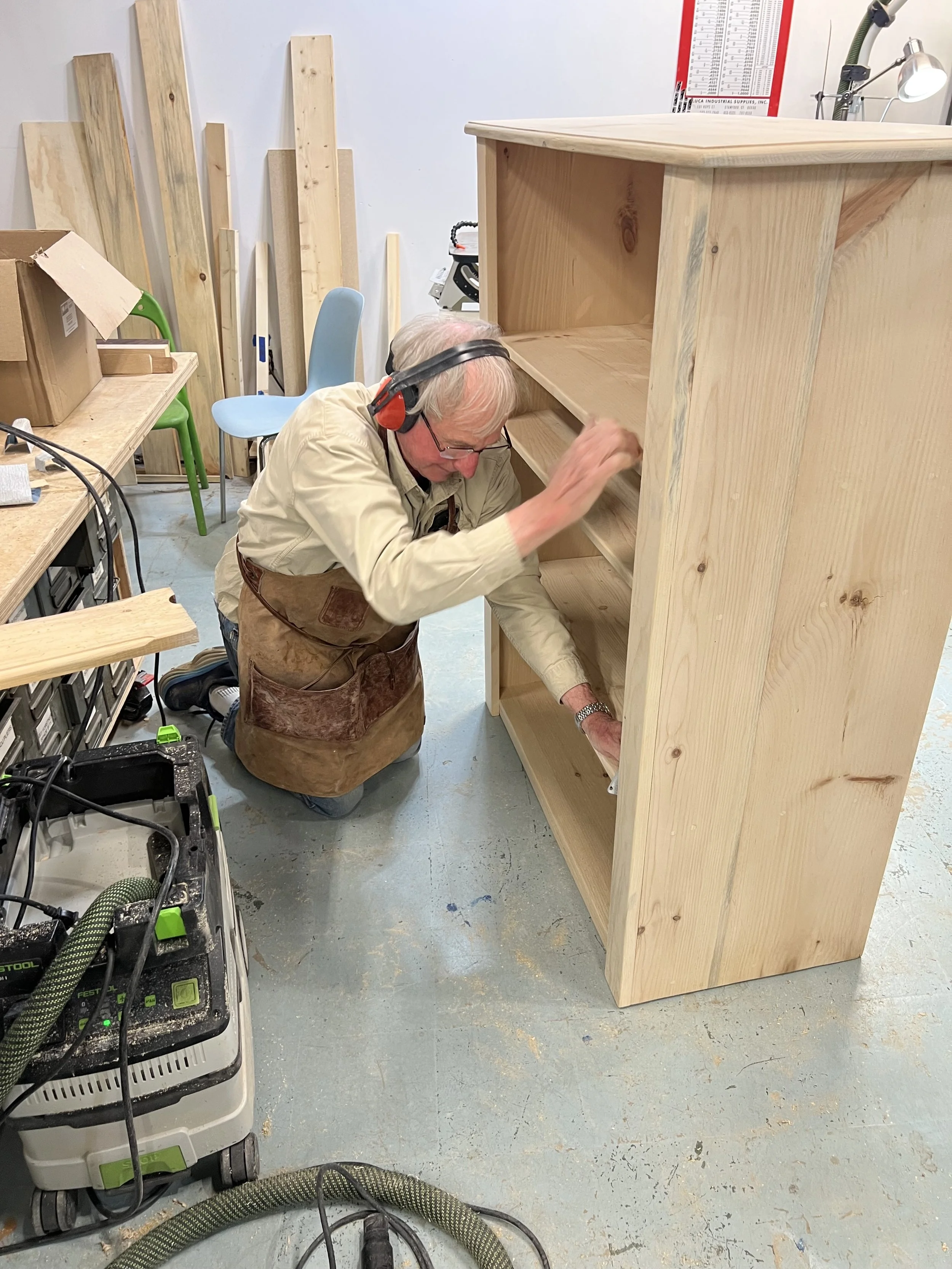 An elderly man working on assembling a wooden bookshelf in a woodworking workshop. He is wearing hearing protection, glasses, a beige shirt, and a brown apron. The workshop has various pieces of lumber against the wall, a blue chair, and several tools and equipment scattered on tables and the floor.