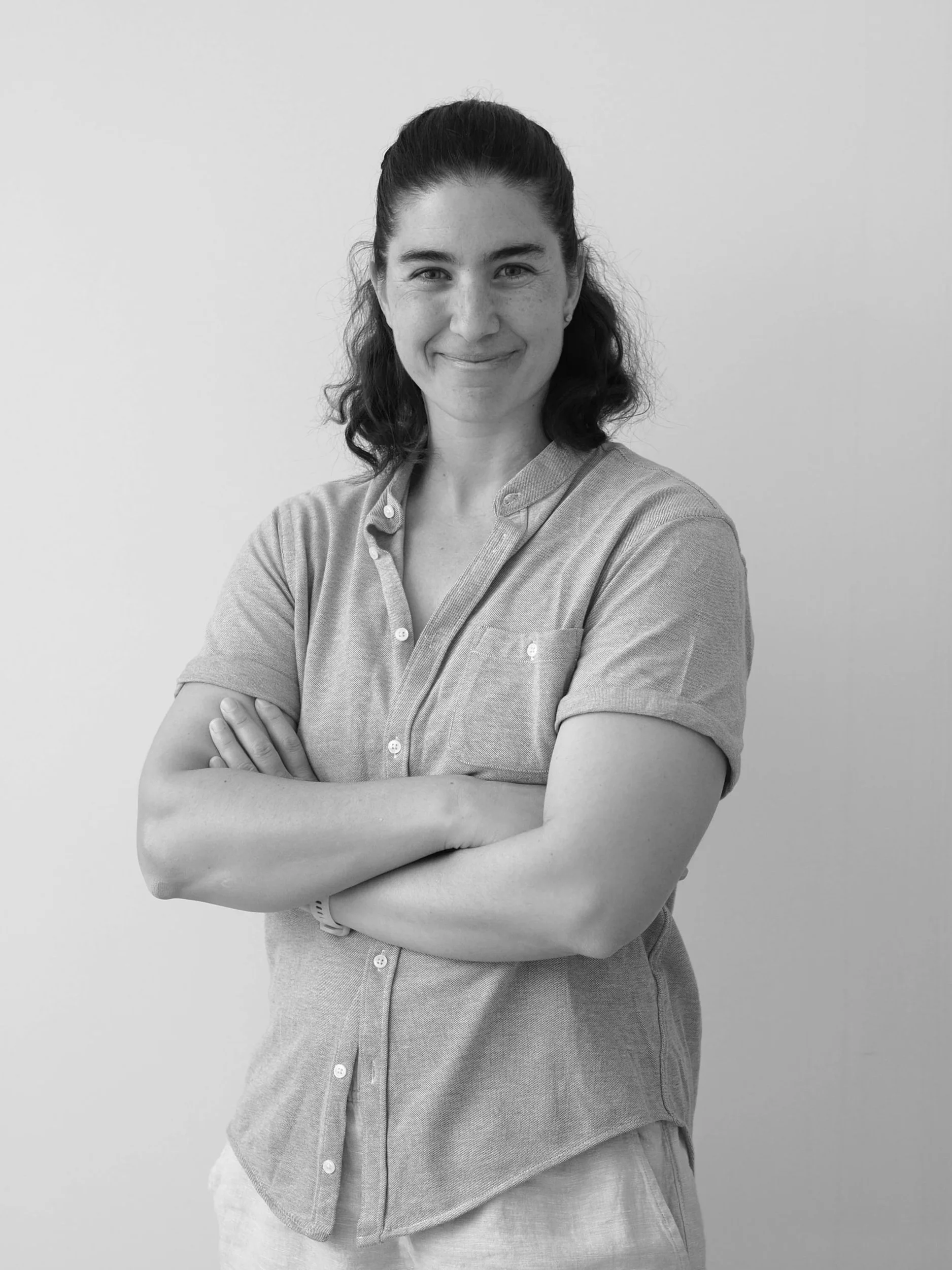 Black and white photograph of a smiling woman with dark, shoulder-length hair, wearing a button-up shirt with the sleeves rolled up, crossed arms, standing against a plain wall.