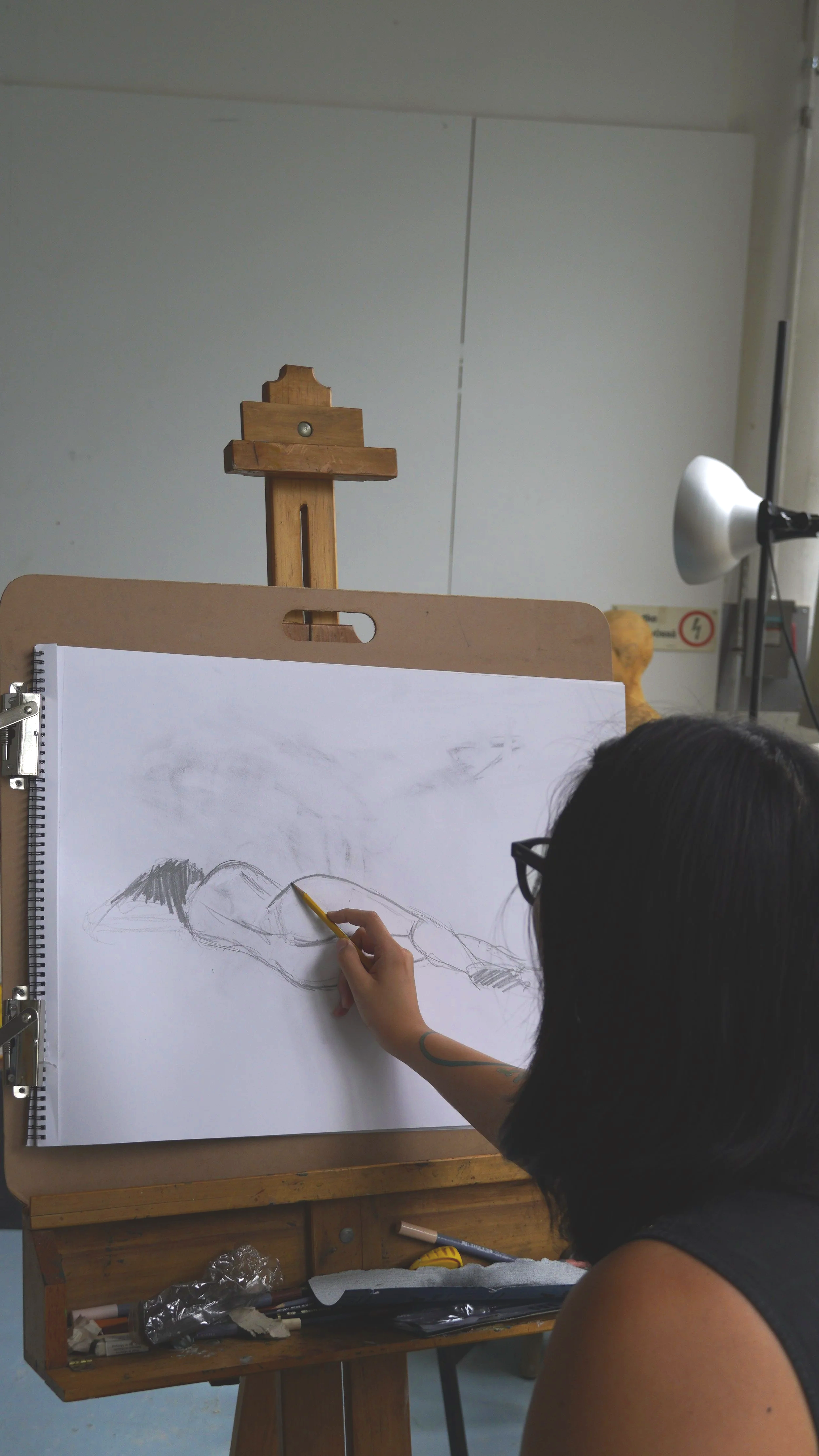 A person drawing a figure on a large white paper attached to a drawing board, with art supplies and a lamp on a wooden table below, an easel and a white wall in the background.