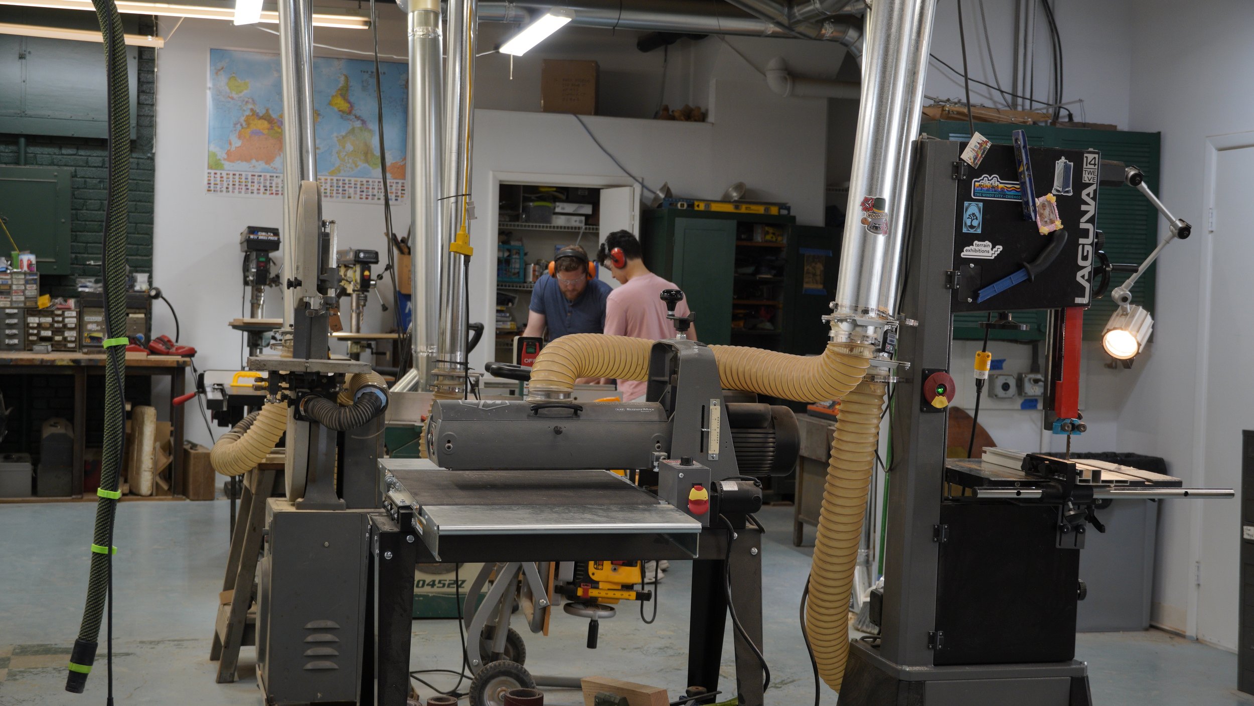 Two people working in a woodworking workshop with various machines and tools, and world maps on the wall.