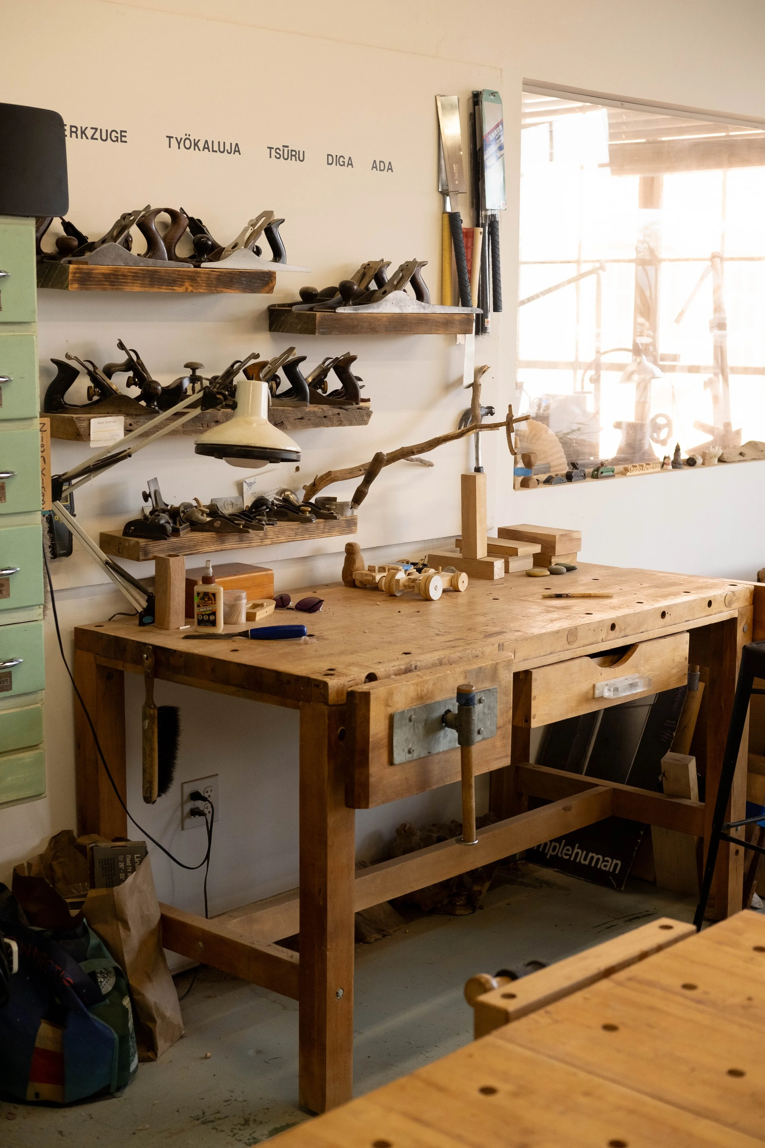 Wooden workbench in a workshop with hand planes, tools, and small wooden projects, and a window in the background.