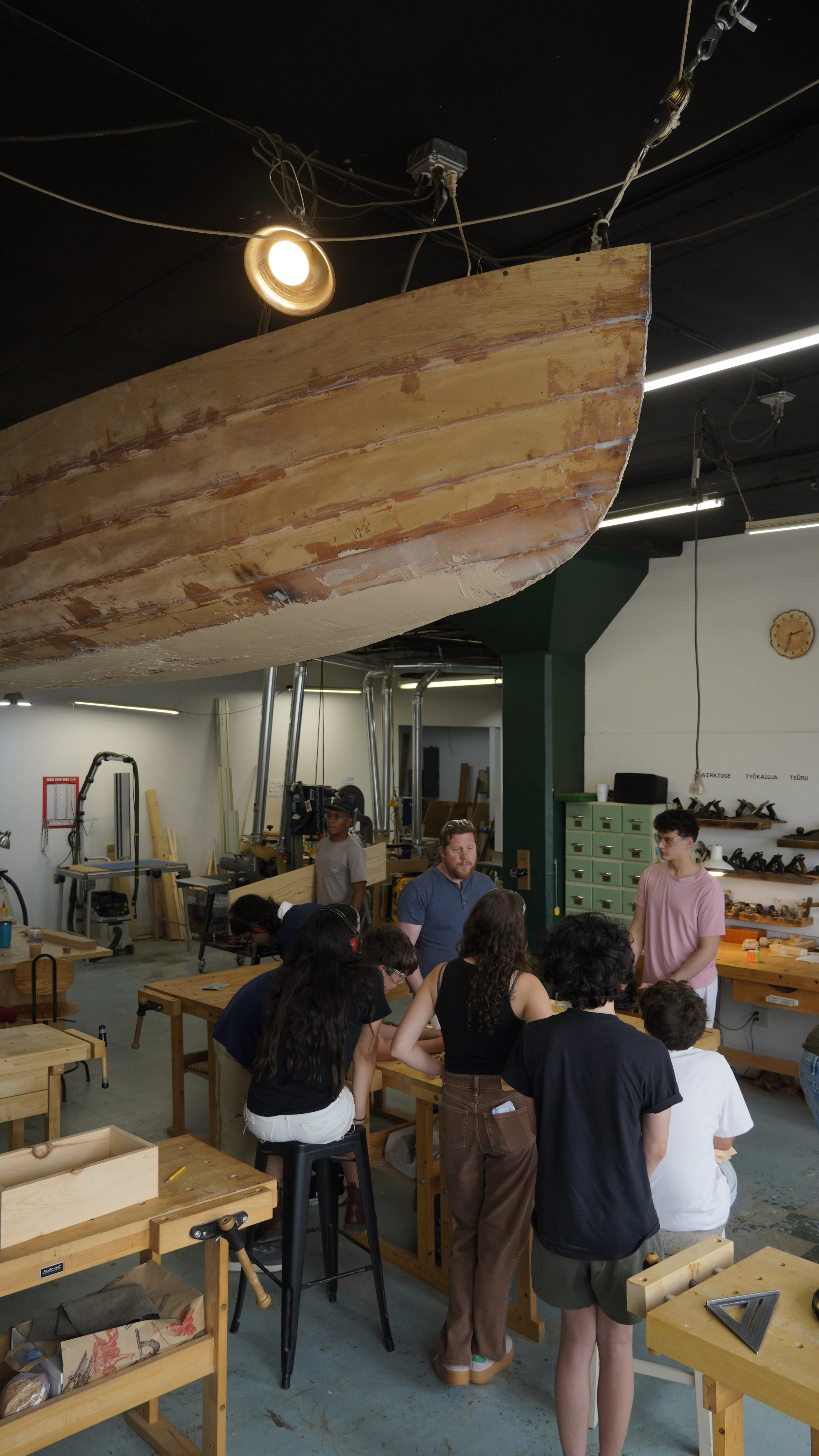 People gathered around wooden tables inside a woodworking workshop, with tools and machines on the walls and in the background, and a large wooden boat suspended from the ceiling.