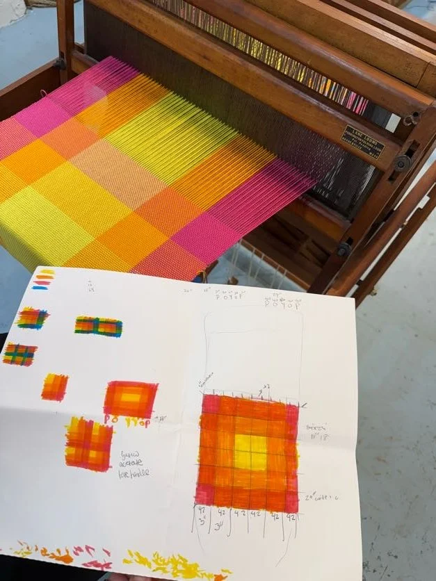 A weaving loom with a colorful woven fabric in pink, yellow, orange, and green. A hand-drawn color chart and pattern diagram on paper are placed in front of the loom, showing color test samples and weaving measurements.