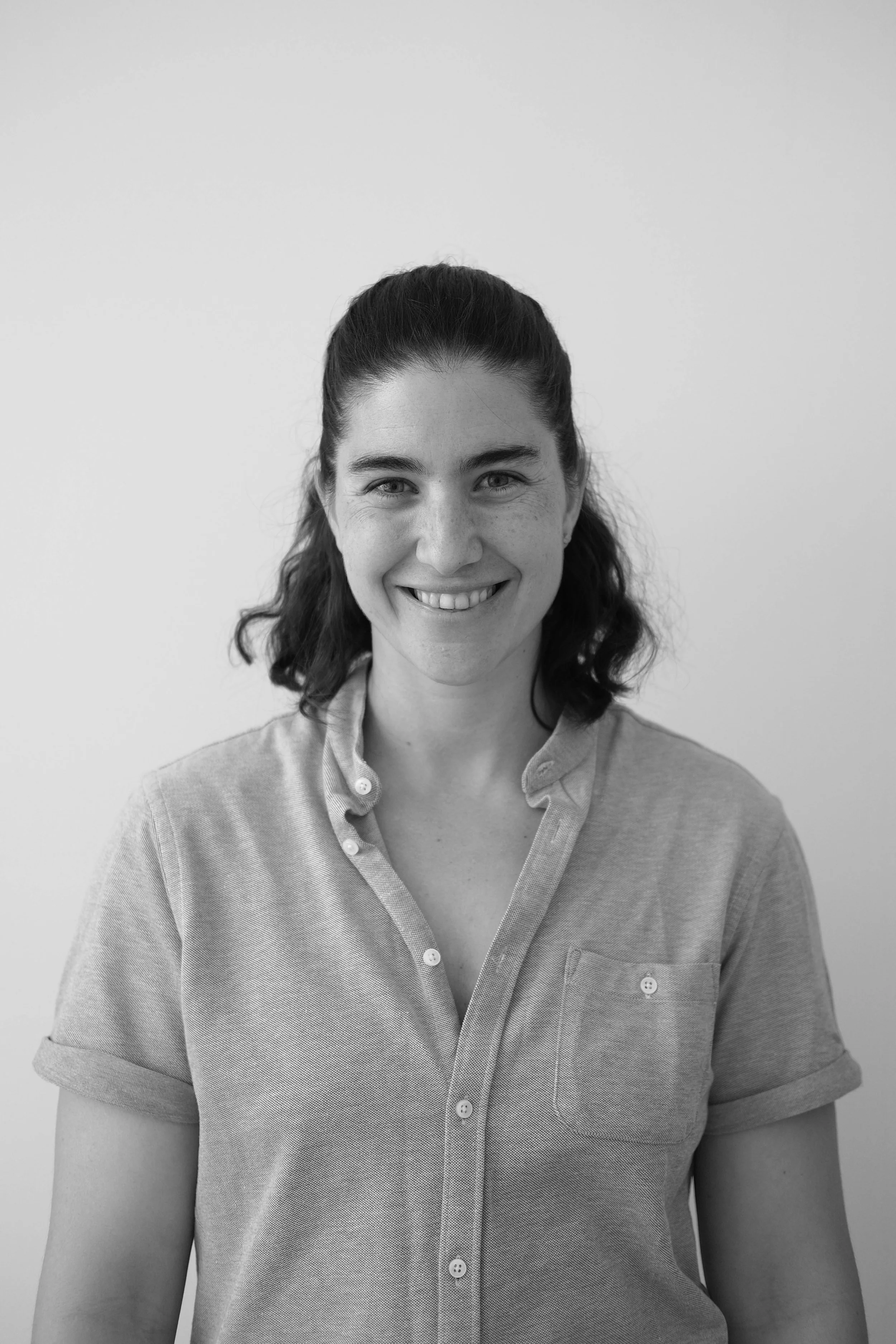Black and white photo of a smiling woman with dark, wavy hair tied back, wearing a casual button-up shirt, standing against a plain background.