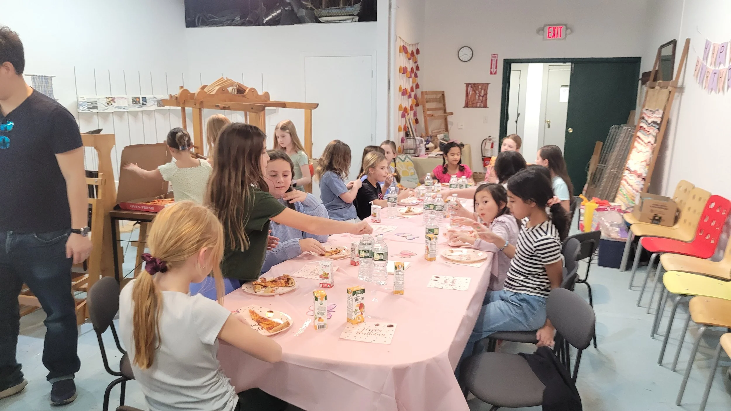 A group of children sitting at a long table having pizza and drinks during a birthday party or celebration in a decorated indoor space.