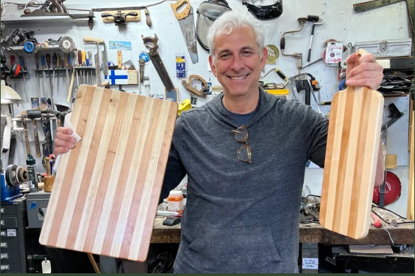 Man smiling in a woodworking workshop holding two wooden cutting boards.