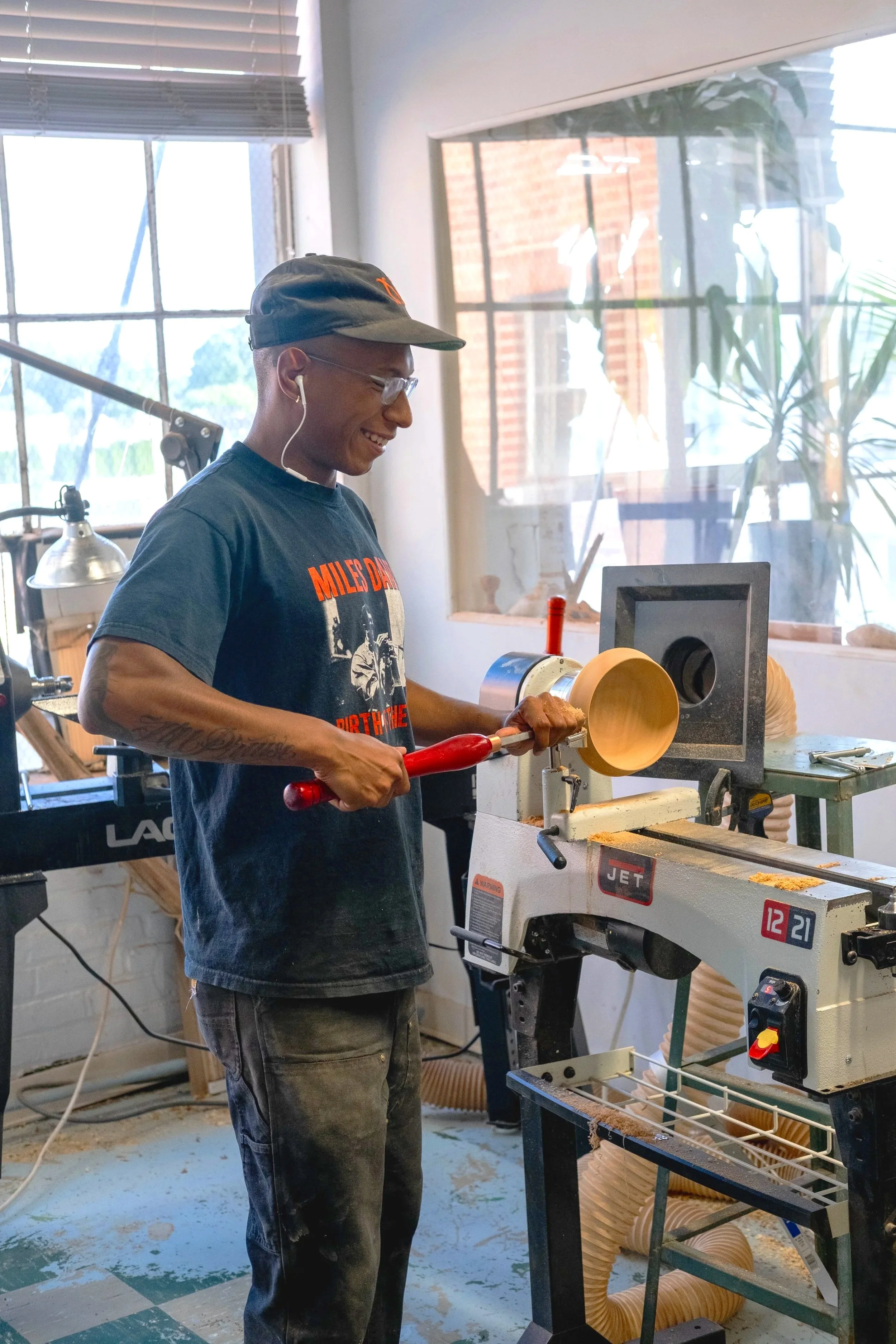 A man wearing glasses, a black cap, and a t-shirt printing a wooden bowl on a woodworking lathe in a workshop with large windows, plants, and woodworking equipment.