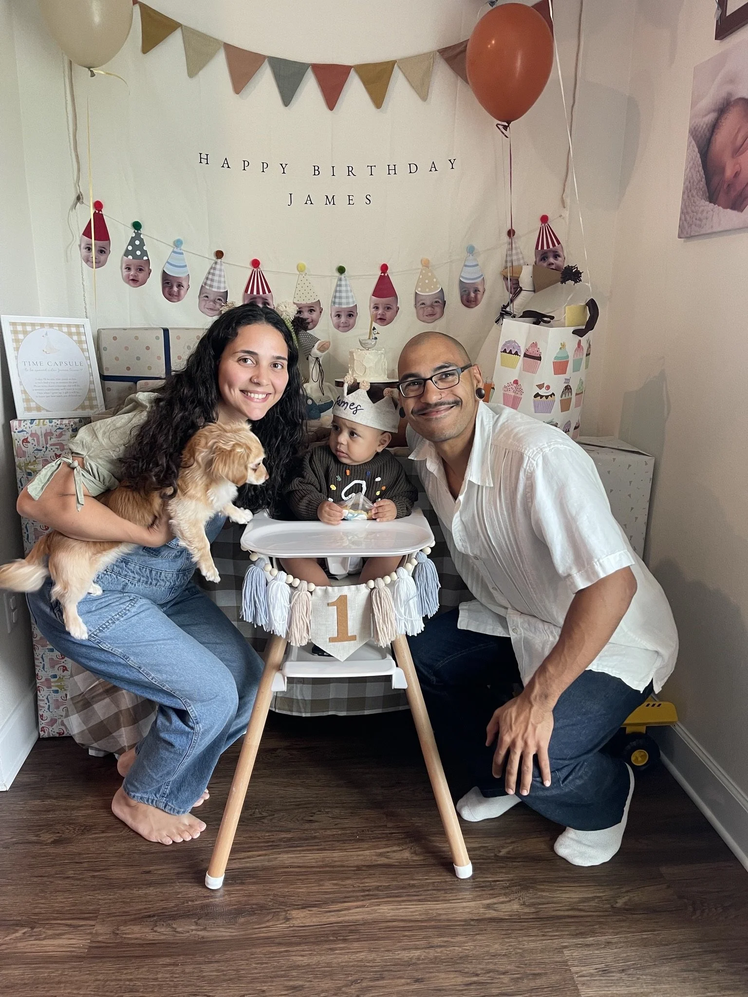 Family celebrating child's first birthday with cake, balloons, and decorations, including a banner and photos. The child is seated in a high chair with a white banner and the number 1 on it, wearing a party hat, and is being held by a woman. A man and dog are also present in the scene.
