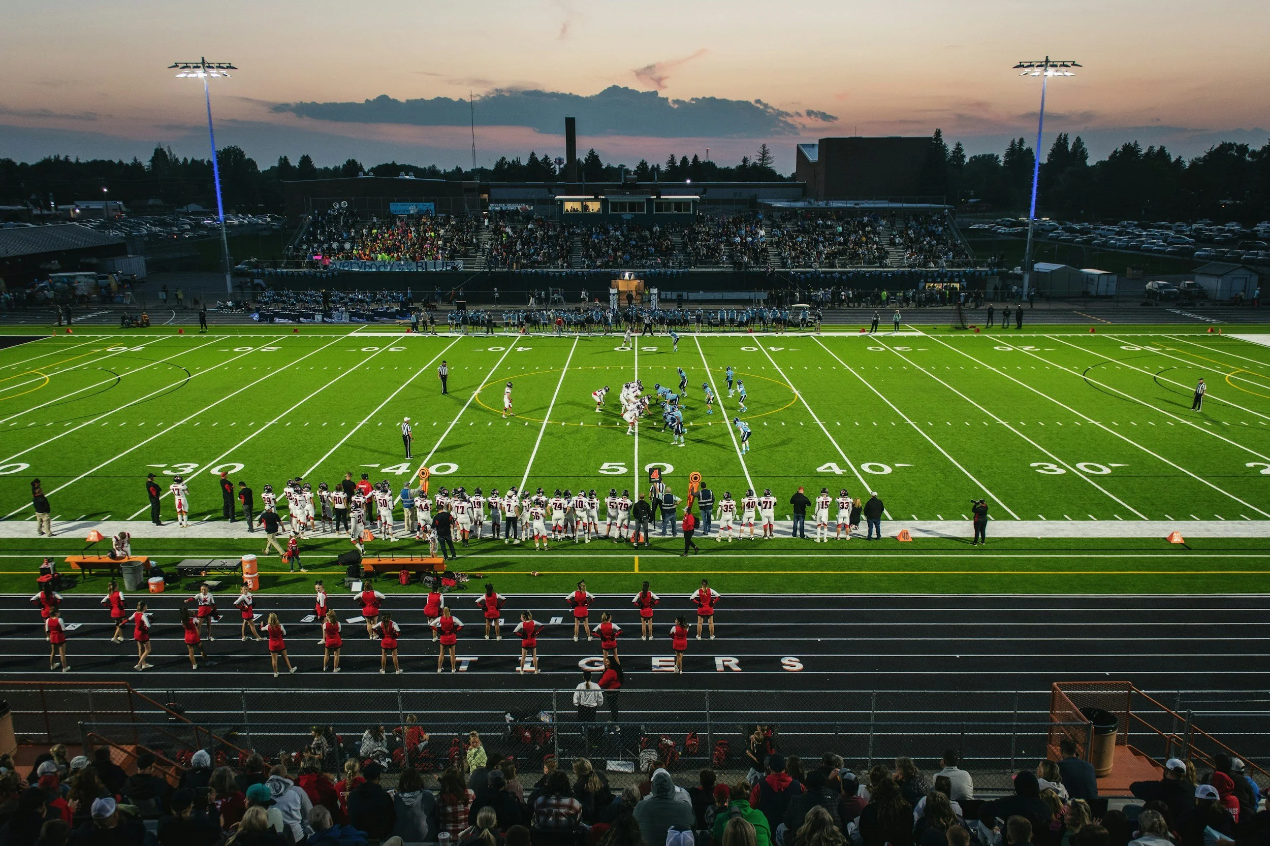 A high school football game at dusk with players on the field, cheerleaders on the sidelines, and a large crowd in the stands under the evening sky.
