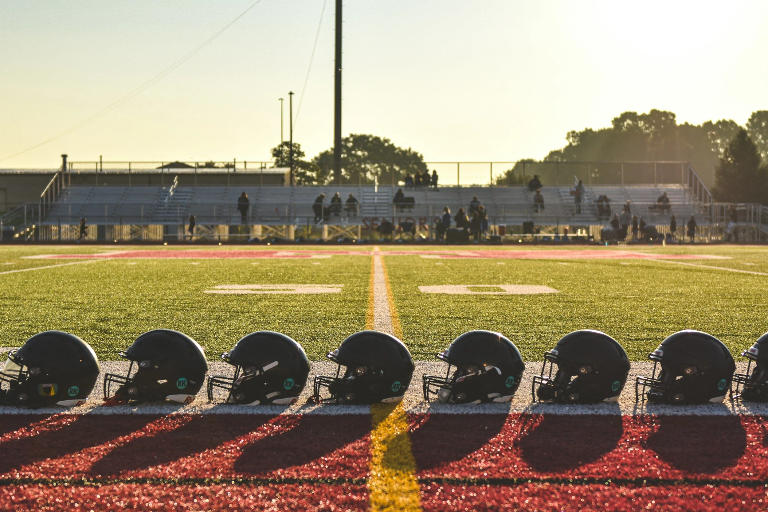 Football helmets lined up on the sideline of a football field with bleachers and players in the background at sunset.