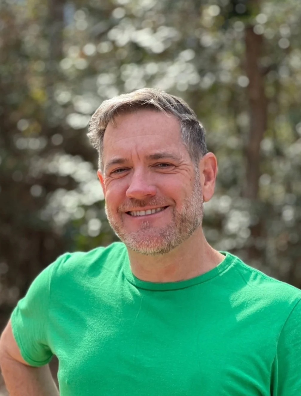A middle-aged man with gray hair and a beard, smiling while standing outdoors in a wooded area, wearing a bright green t-shirt.