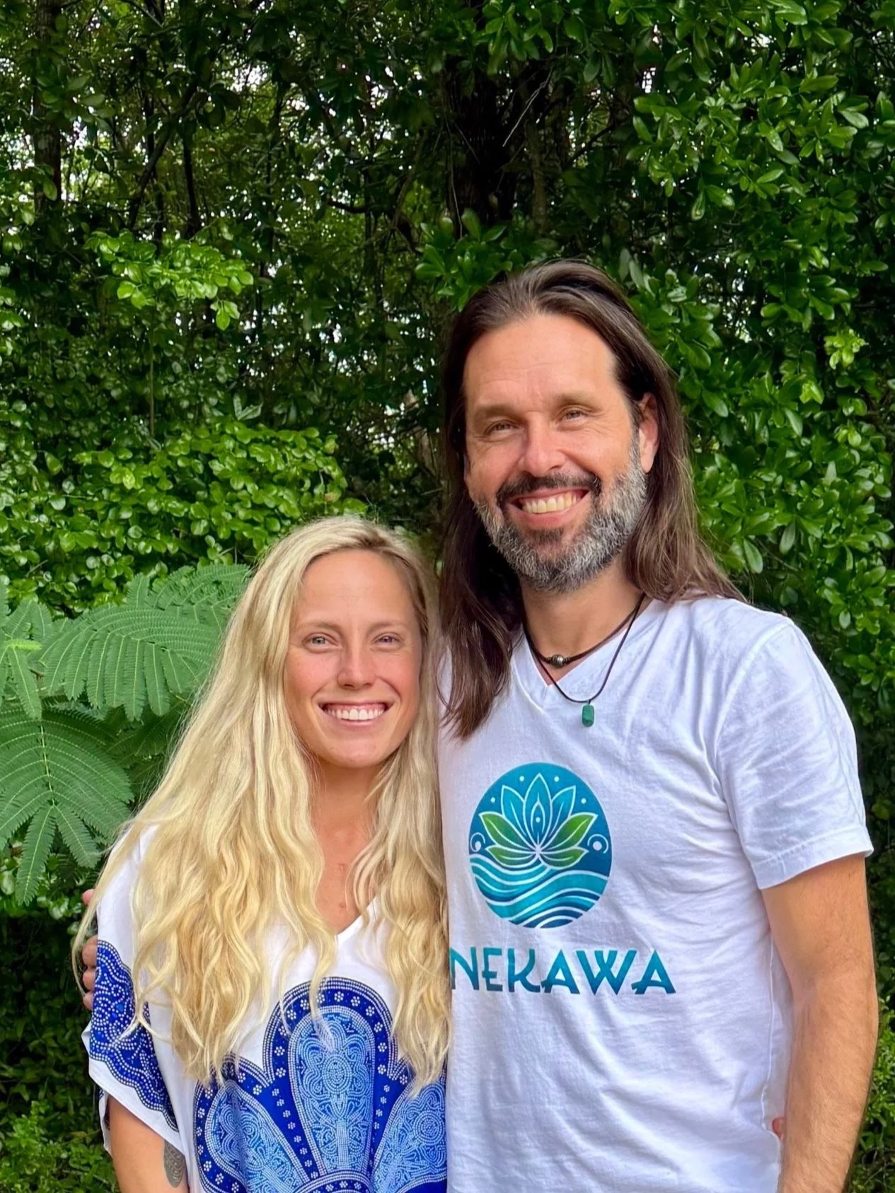 A smiling man and woman standing outdoors in front of green foliage. The woman has long blonde hair and is wearing a white and blue patterned top. The man has long dark hair, a beard, and is wearing a white T-shirt with a blue and green logo that says 'NEKAWA' and features a stylized lotus flower over water.