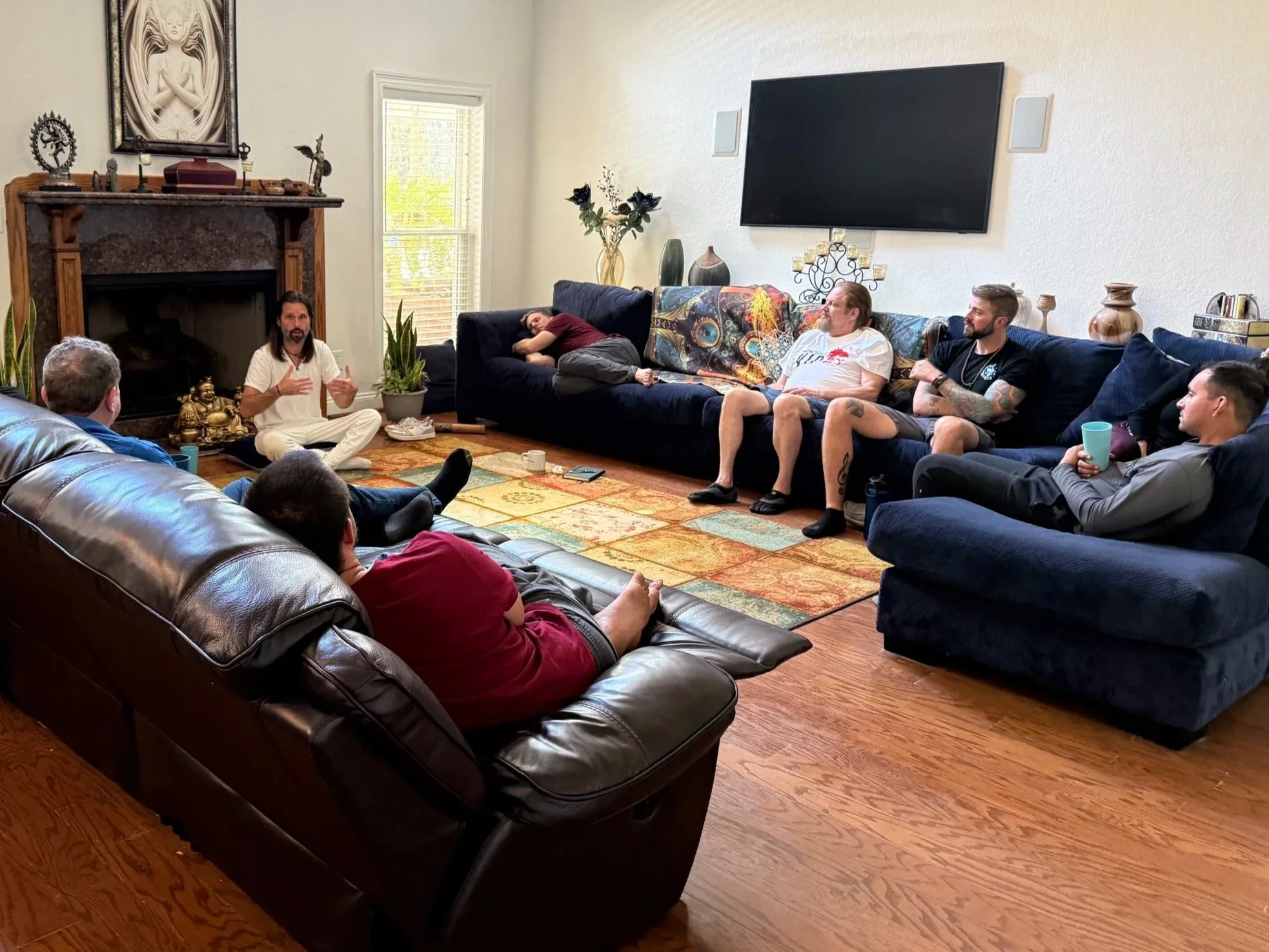 A group of people sitting in a living room, listening to a person speaking near a fireplace. Some are sitting on sofas, one person is lying on the sofa, and another on a leather chair. The room has a large TV on the wall, plants, and decorative items.