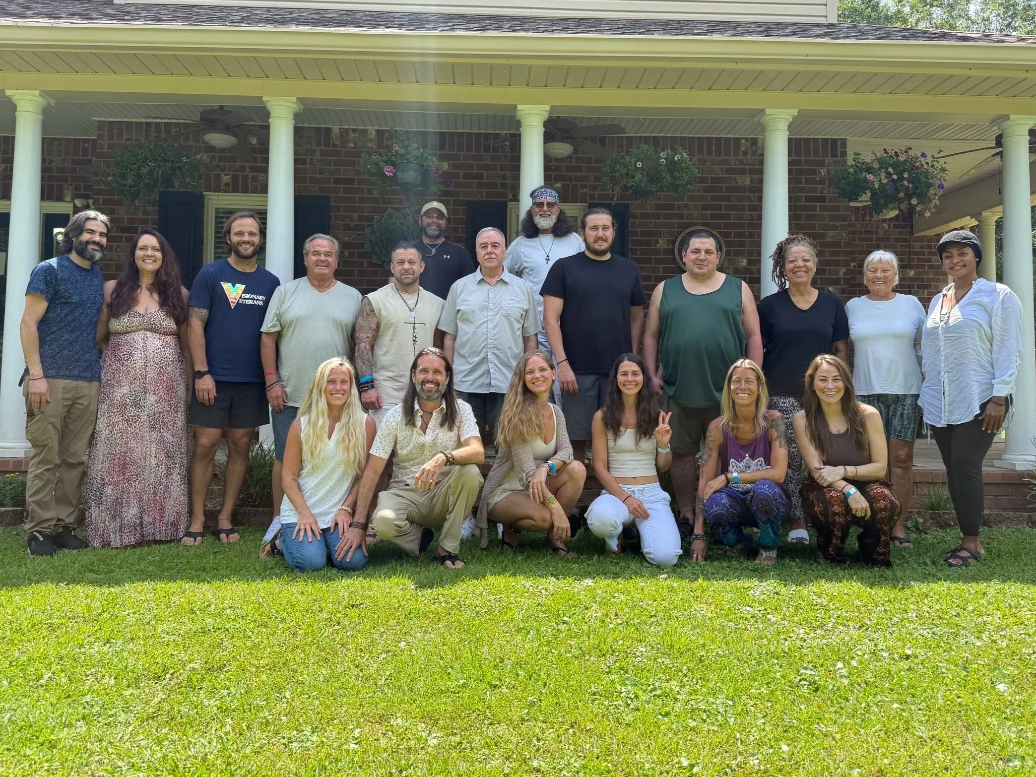 Group of 20 people standing and kneeling on a lawn in front of a house with brick walls and white columns, smiling for a photo.