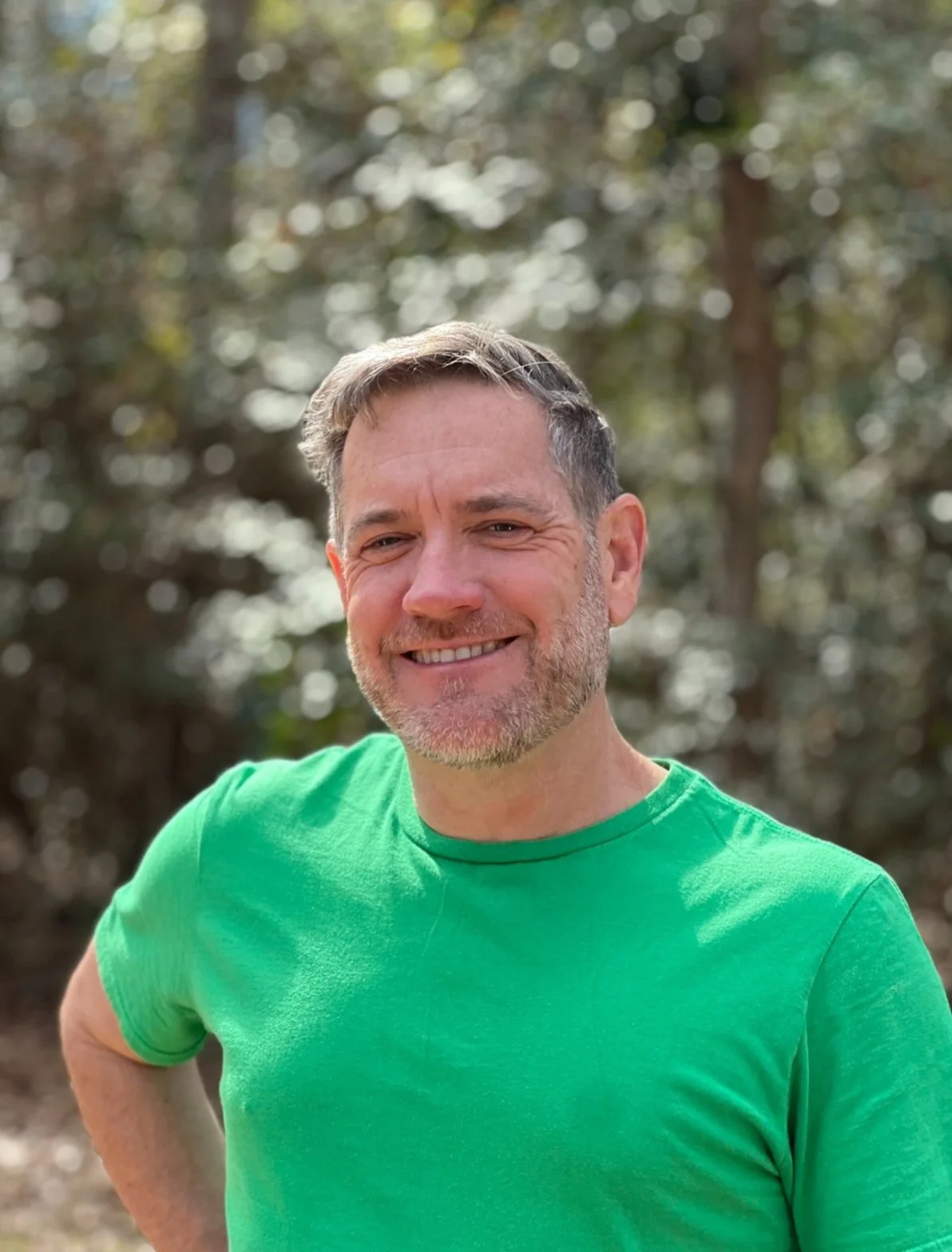 A smiling man in a green T-shirt outdoors with blurred trees in the background.