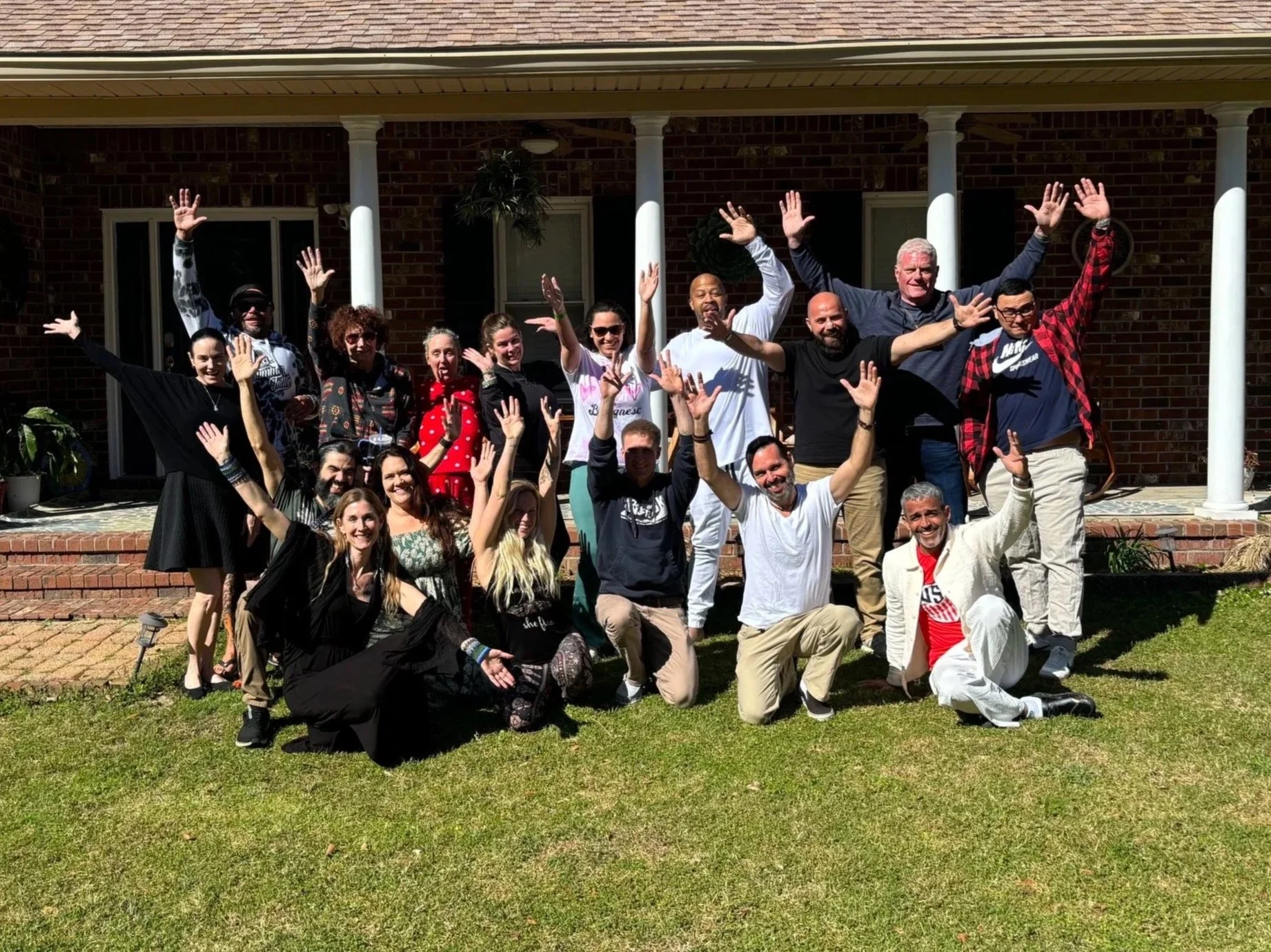 Group of people standing and kneeling on a lawn in front of a brick house, smiling and raising their hands in celebration.